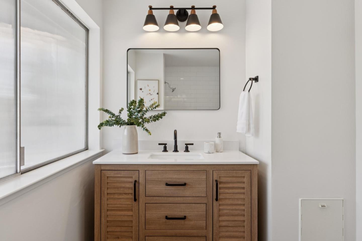 Bathroom vanity with a wood cabinet, black faucet, rectangular mirror, and greenery beside a large frosted window.