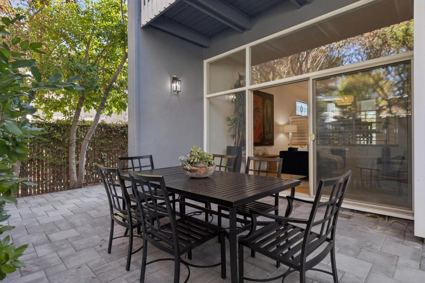 Outdoor dining patio with gray pavers, black furniture, and sliding glass doors beside leafy trees.