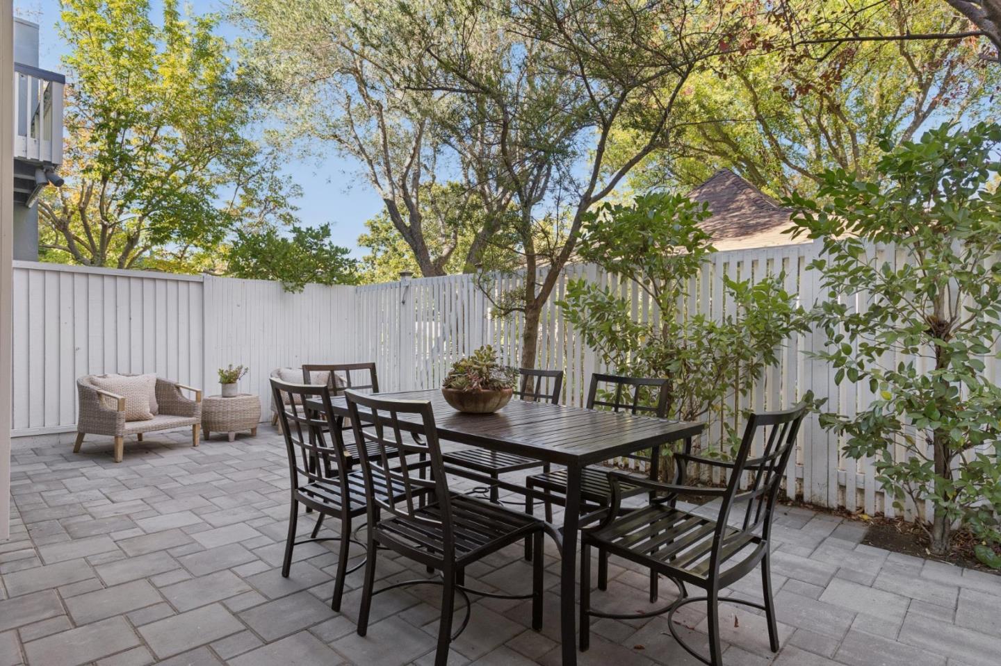 Fenced patio with a dark dining set, wicker lounge chair, and leafy trees.