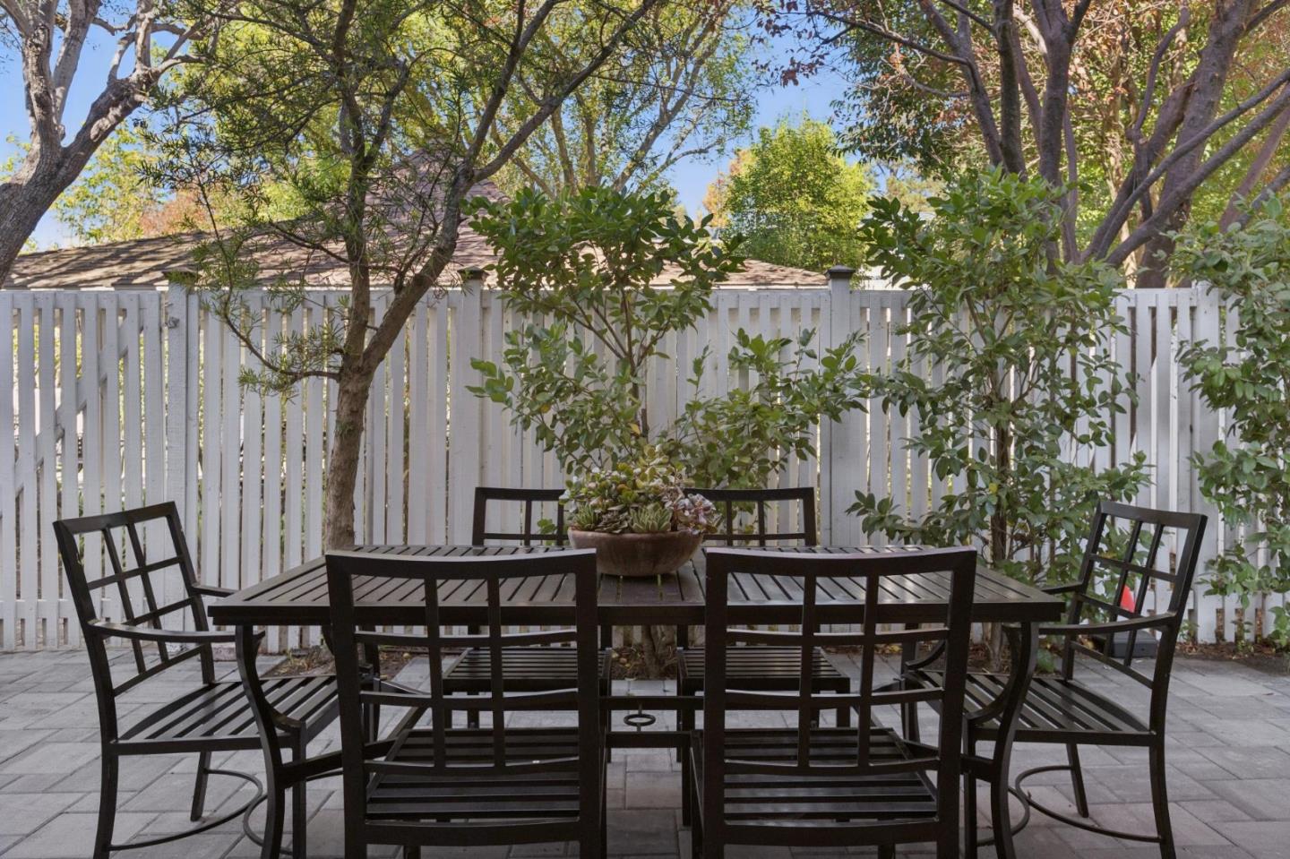 Outdoor patio with black dining set, gray pavers, white fence, and mature trees.