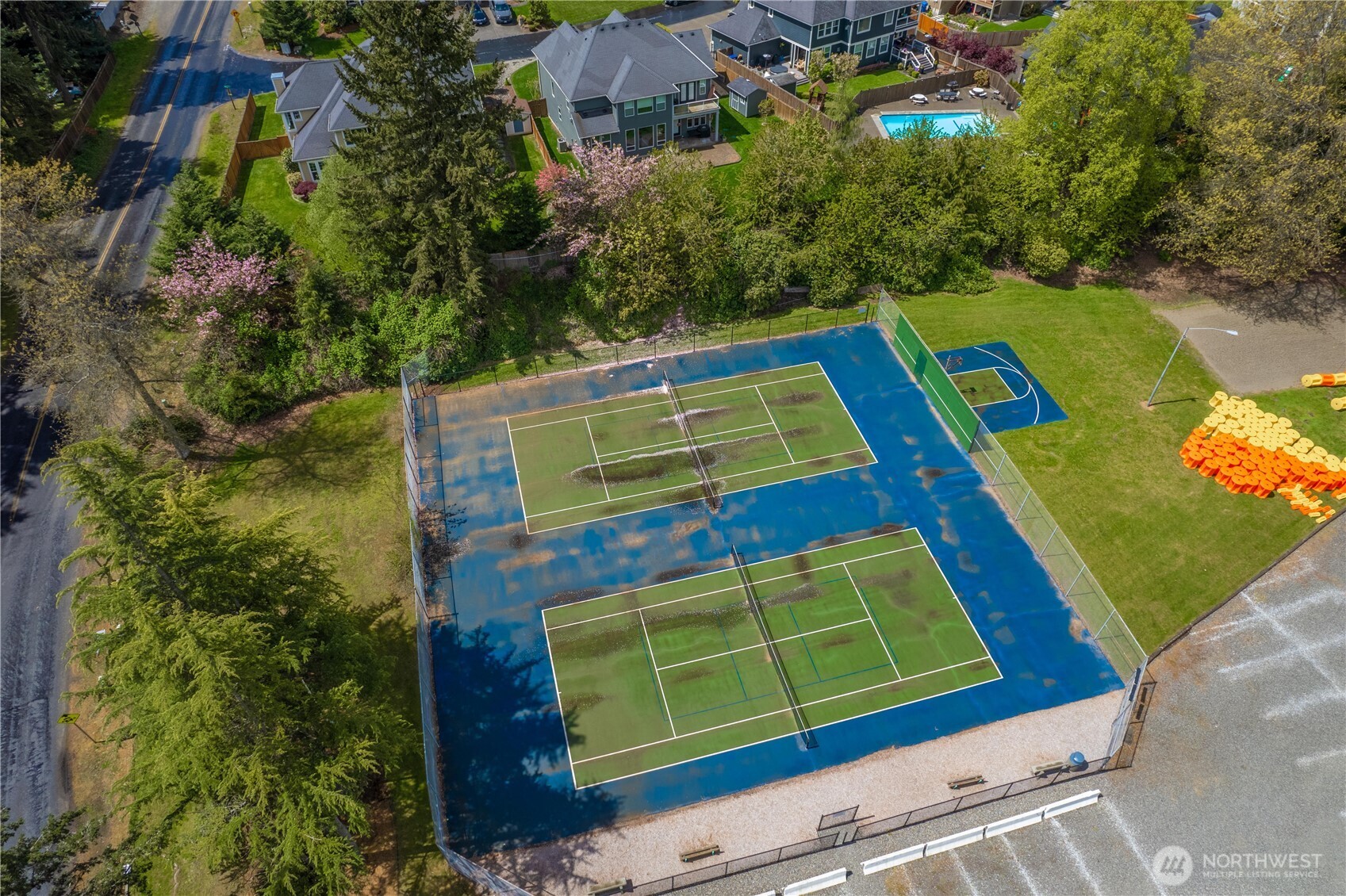 Aerial view of two fenced tennis courts beside a basketball court and neighboring homes.