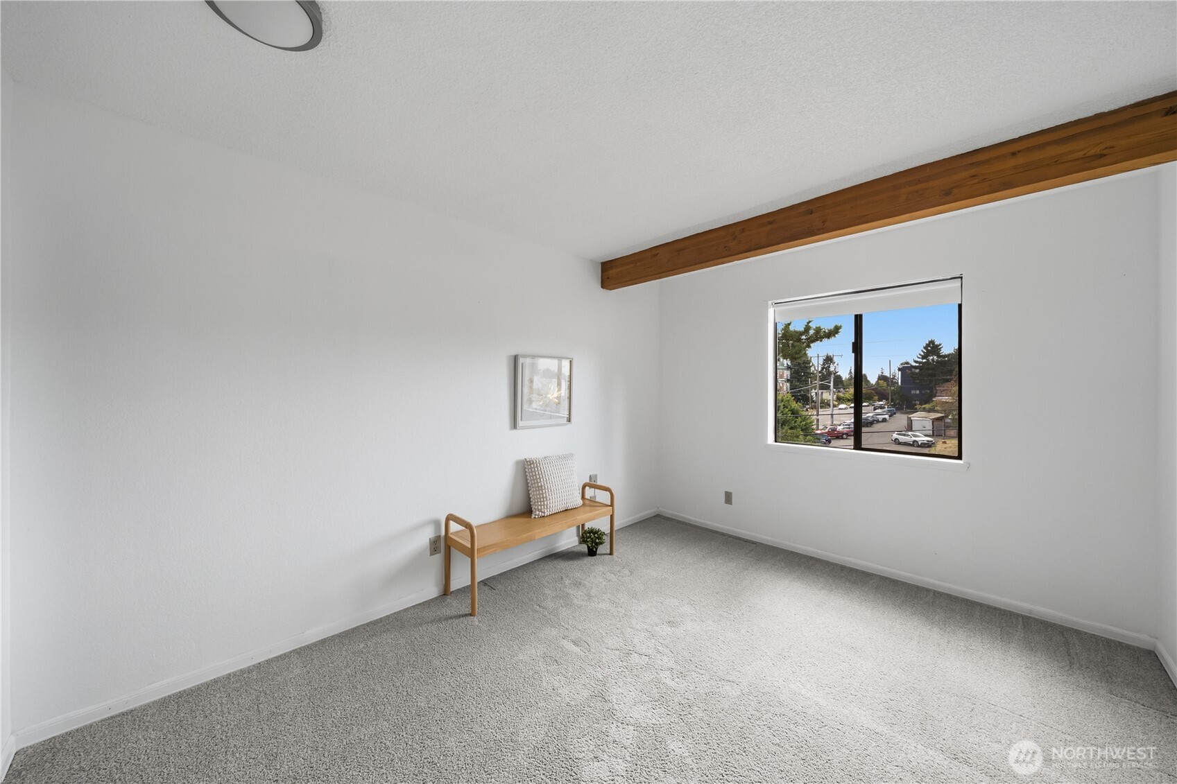 Bedroom with gray carpet, an exposed wood beam, a wooden bench, and a window overlooking a parking area.