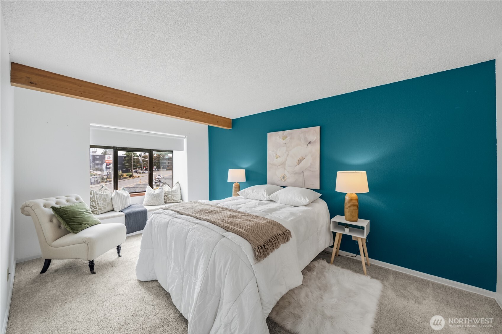 Staged bedroom with a white bed, teal accent wall, exposed wood beam, and cushioned window seat.