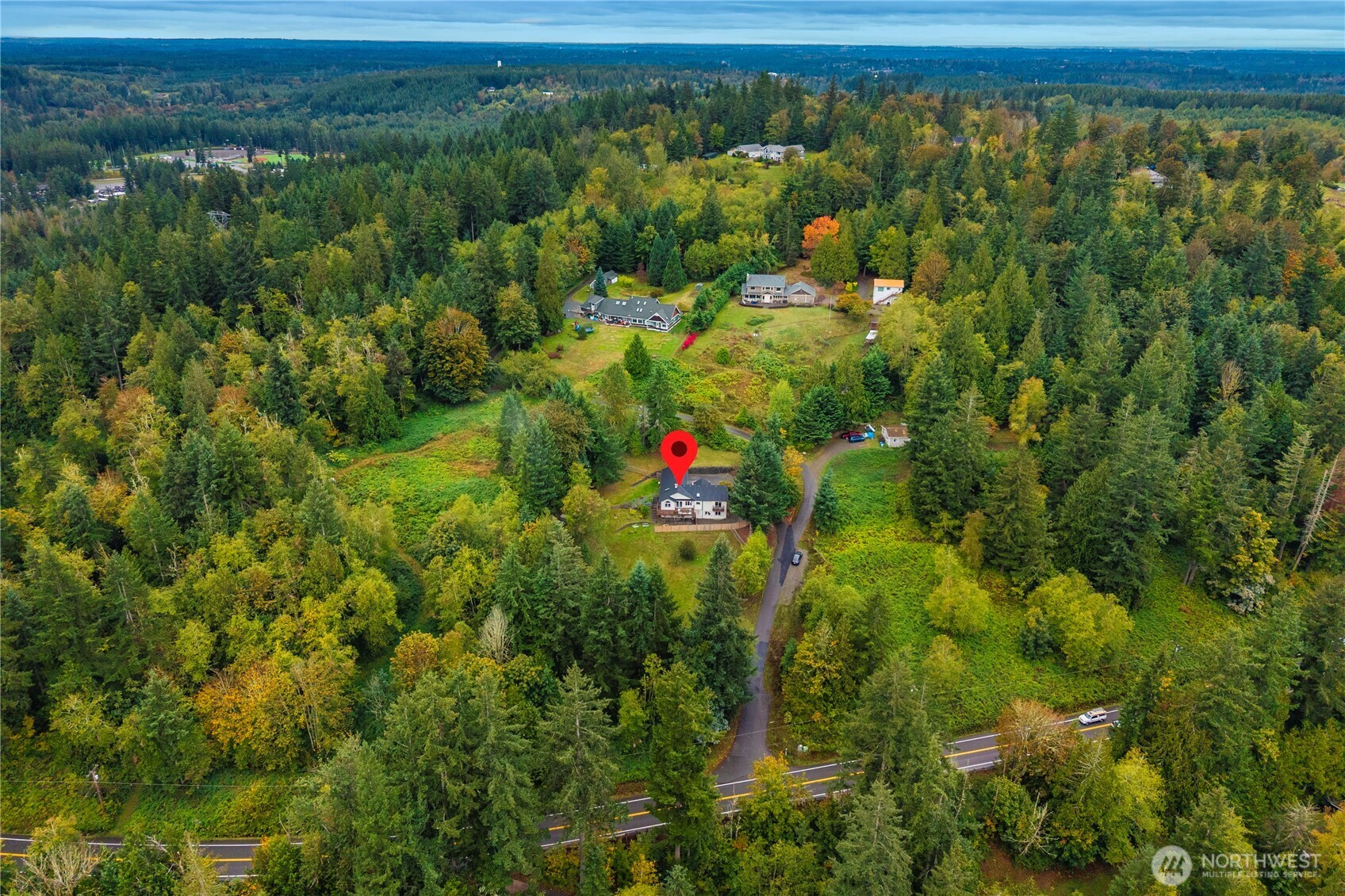 Aerial view of a wooded rural home marked by a red pin, with neighboring homes and a road nearby.