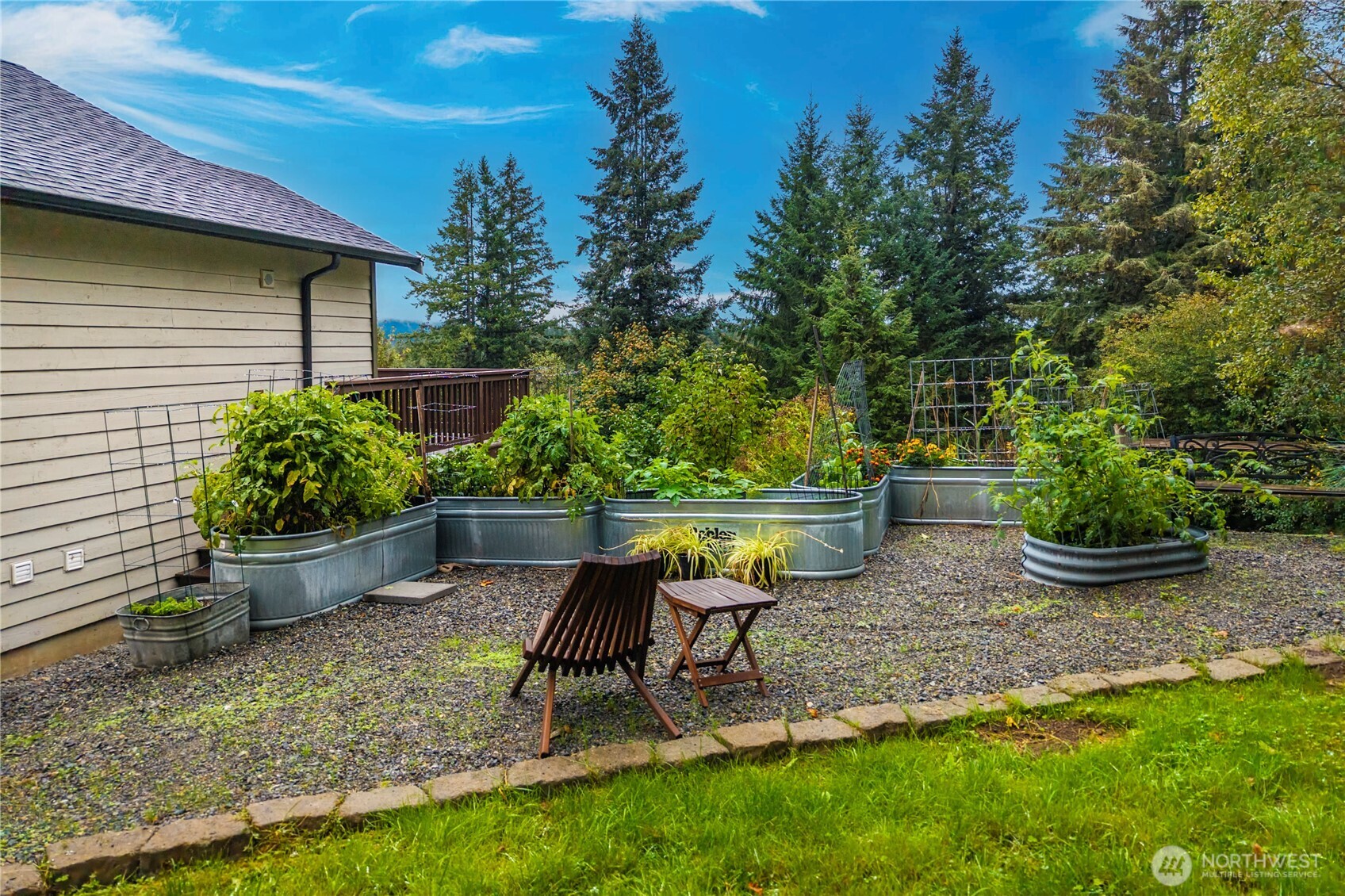 Gravel garden with galvanized raised beds, trellises, and wooden chairs beside a house.