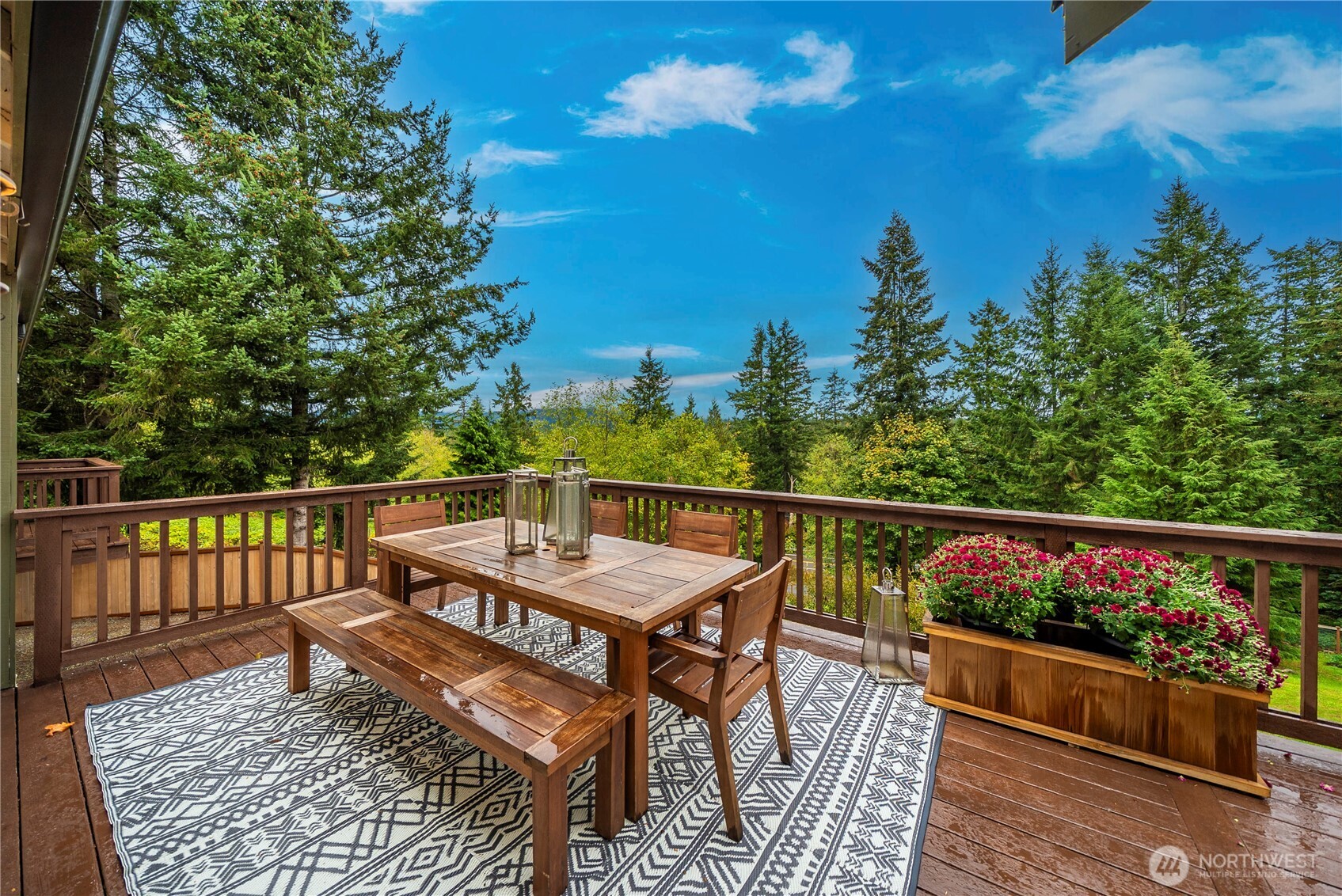 Wooden deck dining area overlooking evergreen trees, with a long table, bench, and flower planter.