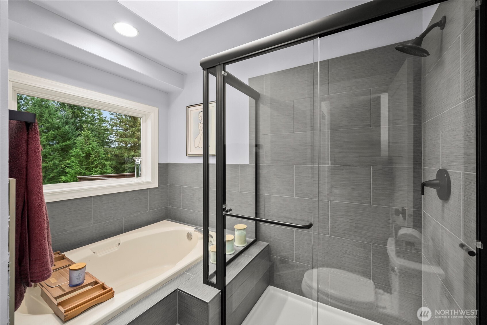 Bathroom with a soaking tub, glass-enclosed shower, gray tile, and a window overlooking evergreens.