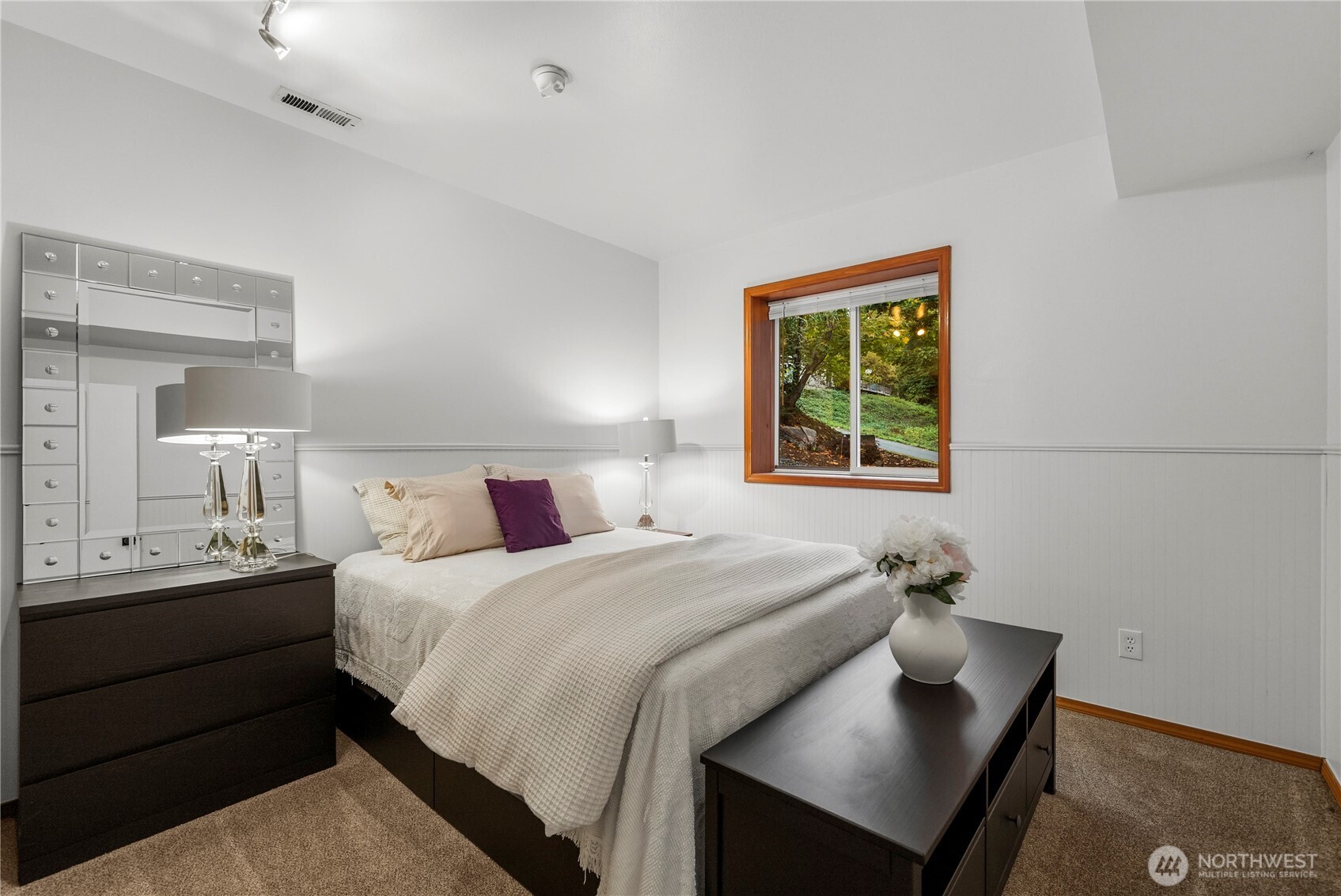 Furnished bedroom with a mirrored headboard and wood-trimmed window overlooking greenery.