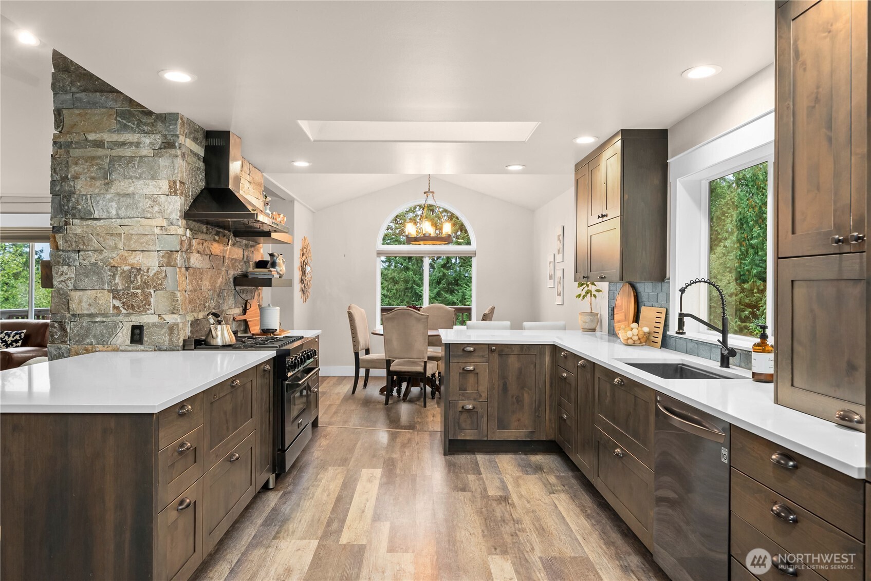 Kitchen with dark wood cabinets, white counters, a stone chimney, and an arched window overlooking trees.