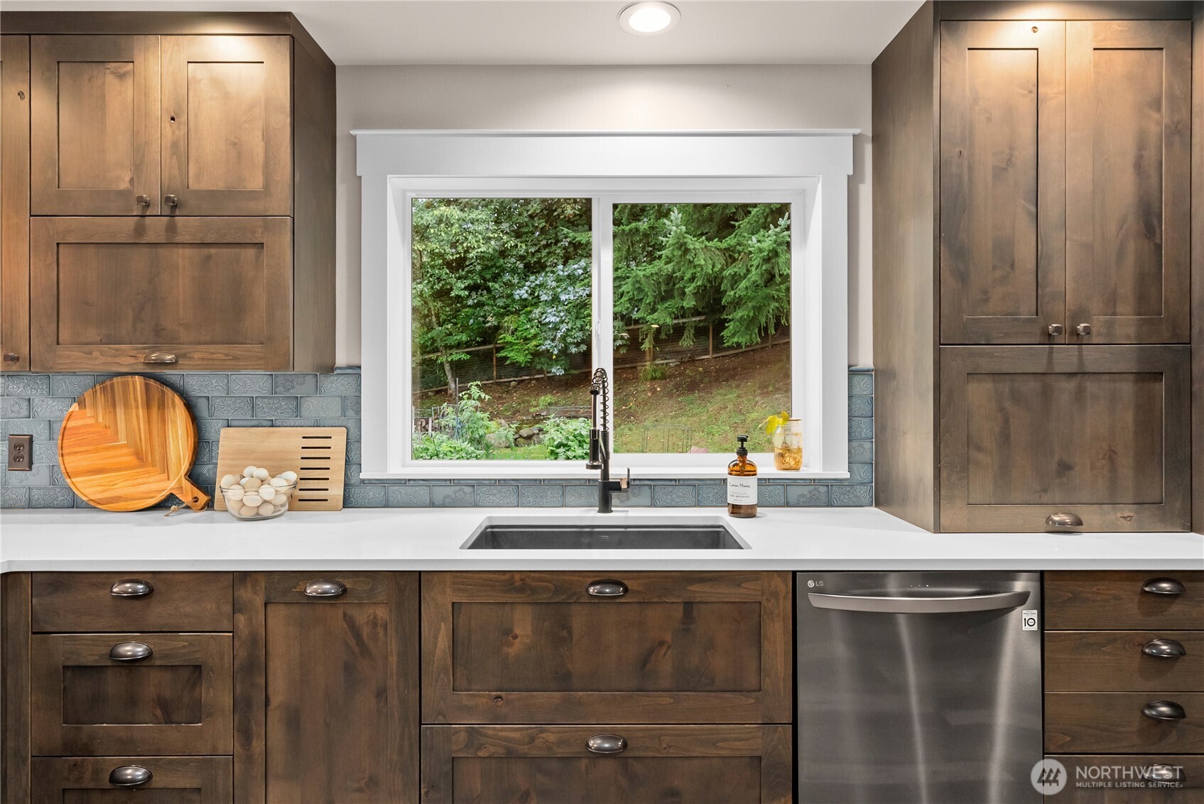 Kitchen with dark wood cabinets, blue tile backsplash, and a sink beneath a window overlooking trees.