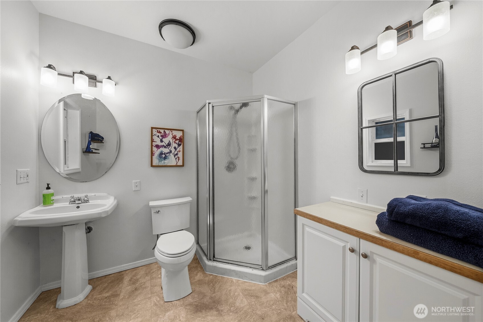 Bathroom with a corner glass shower, pedestal sink, and white vanity topped with folded navy towels.