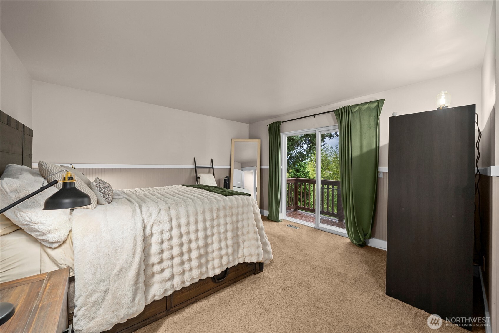 Bedroom with white textured bedding and green curtains beside sliding doors to a tree-lined deck.