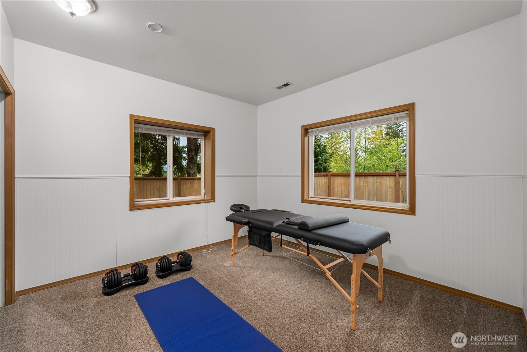 Exercise room with a massage table, adjustable dumbbells, and a blue yoga mat beside two windows.