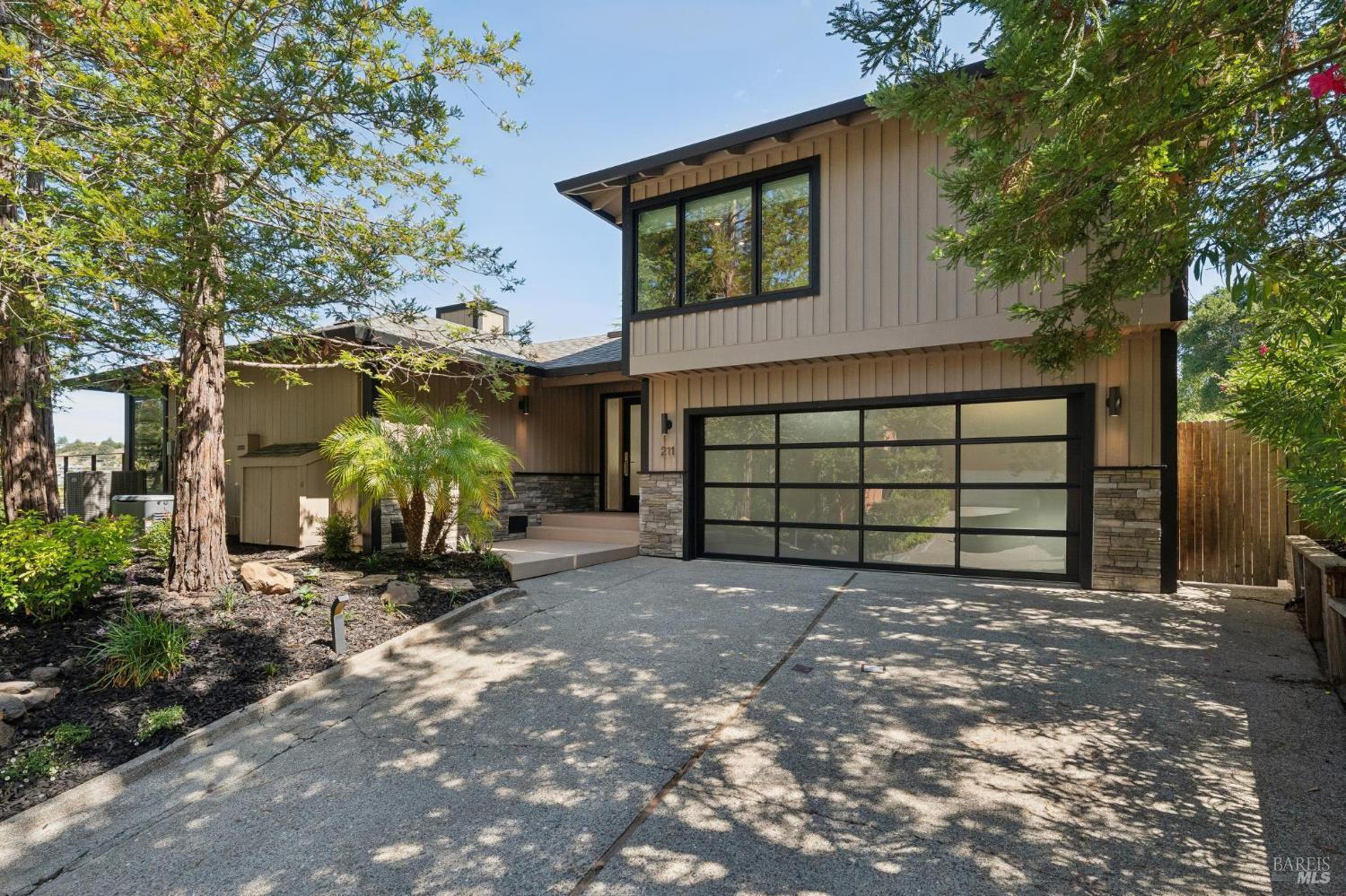 Contemporary two-story home with tan siding, a glass-paneled garage door, and a tree-shaded driveway.