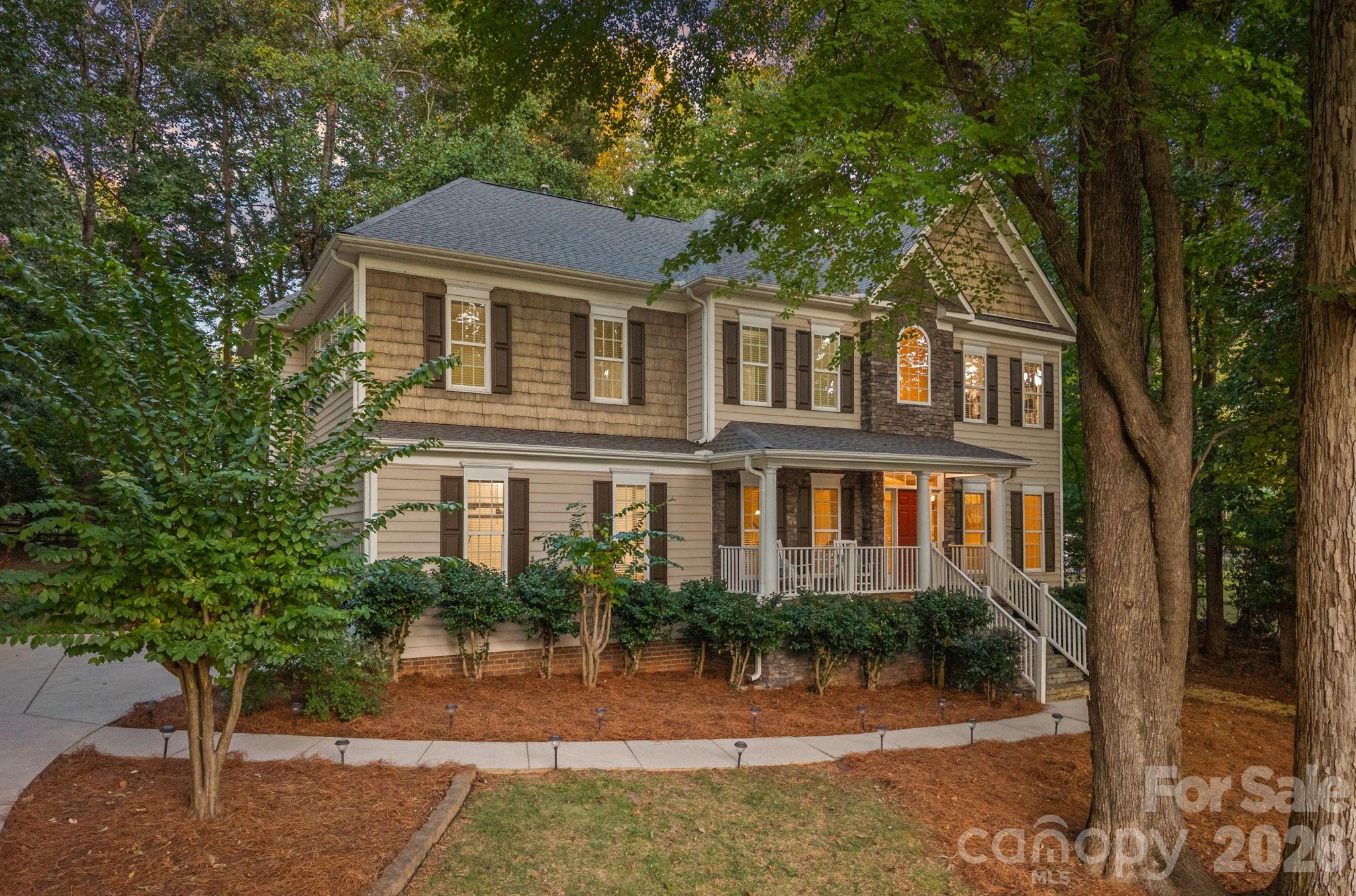 Two-story tan house with stone accents, a covered porch, and mature trees at dusk.