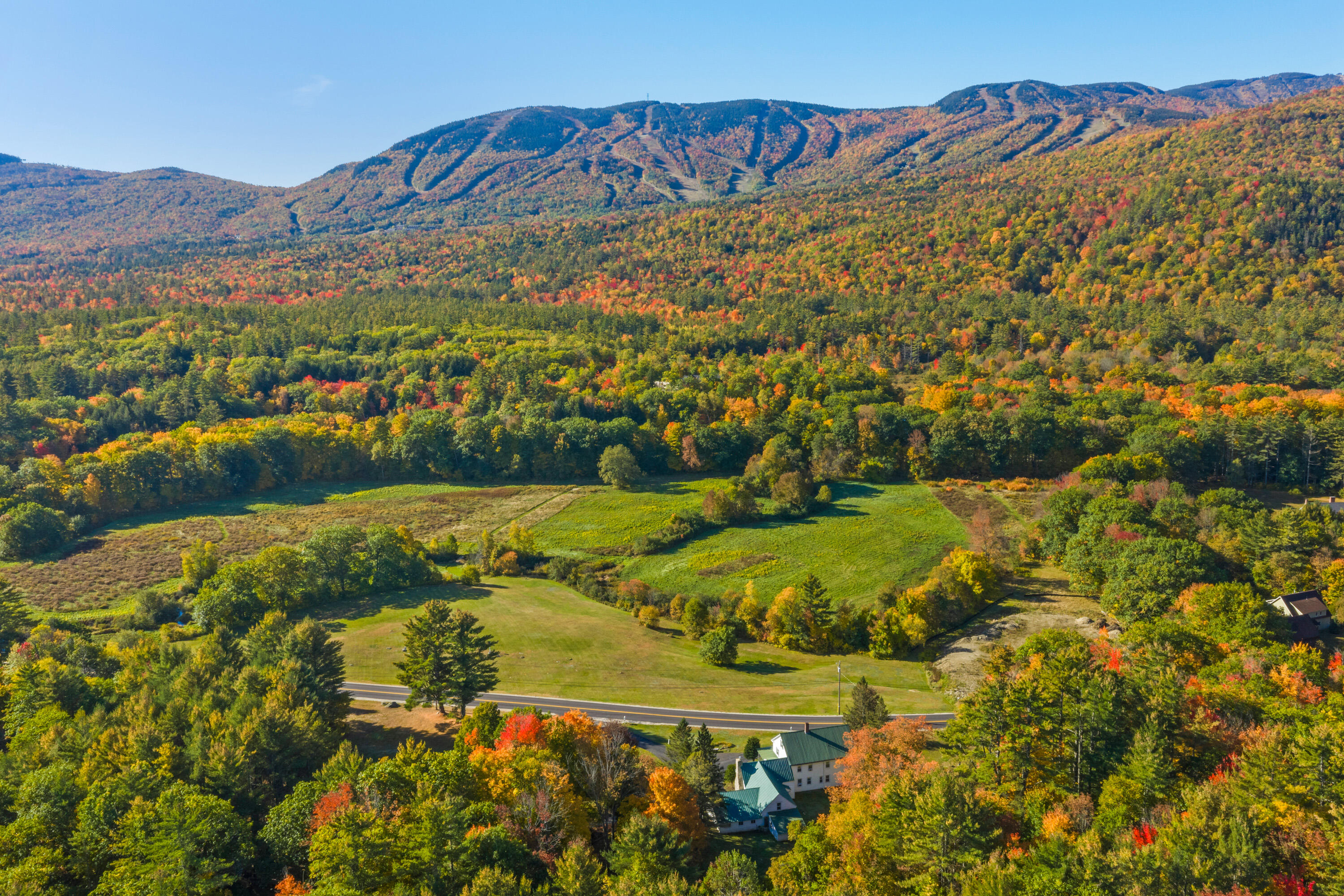 Aerial view of buildings and open grounds amid autumn foliage, with forested mountain slopes beyond.