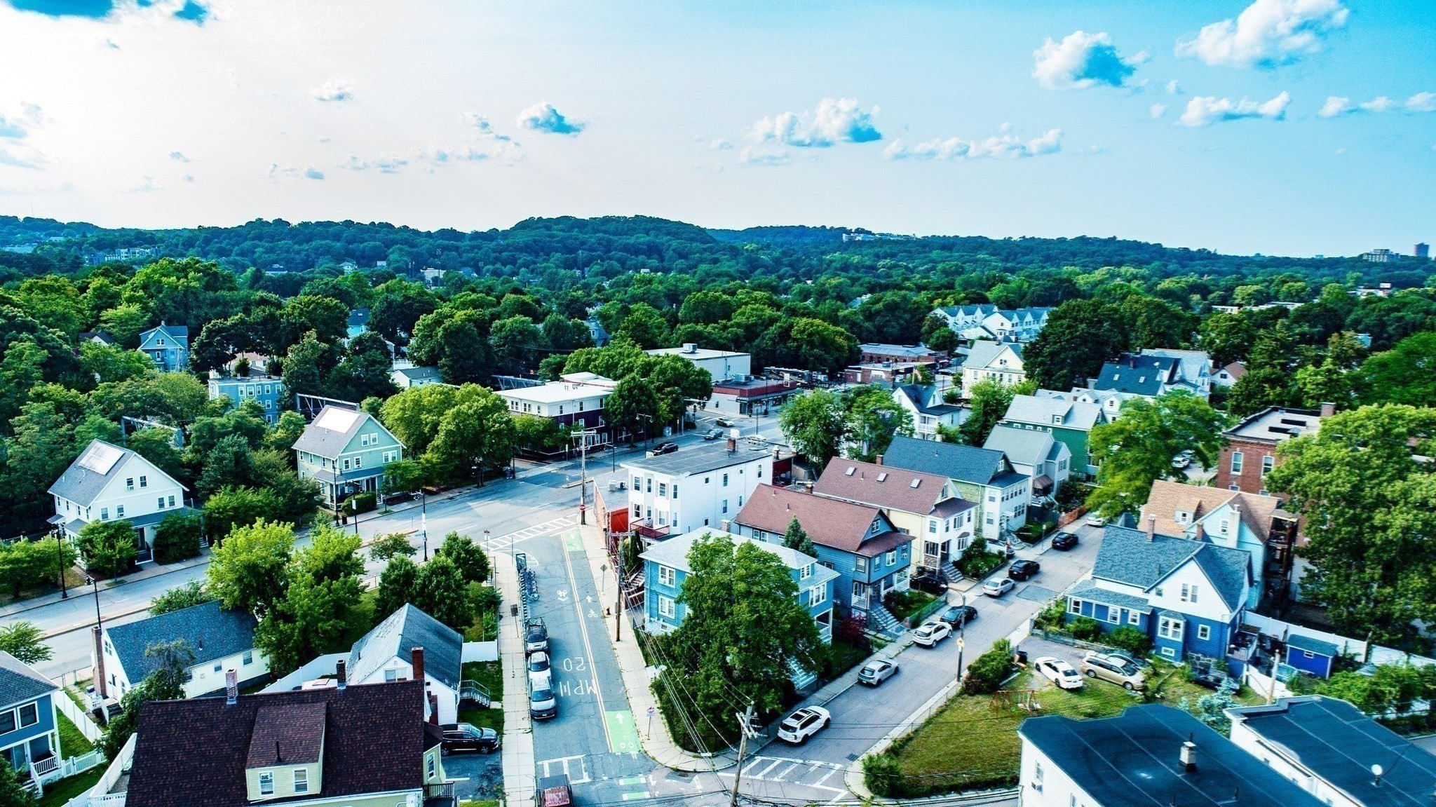 Aerial view of a leafy Boston neighborhood with residential streets and wooded hills beyond.