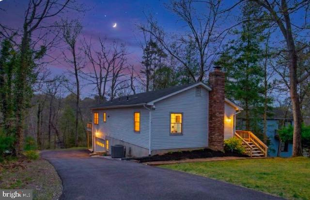 Gray-sided house with a driveway, brick chimney, and illuminated windows at dusk.
