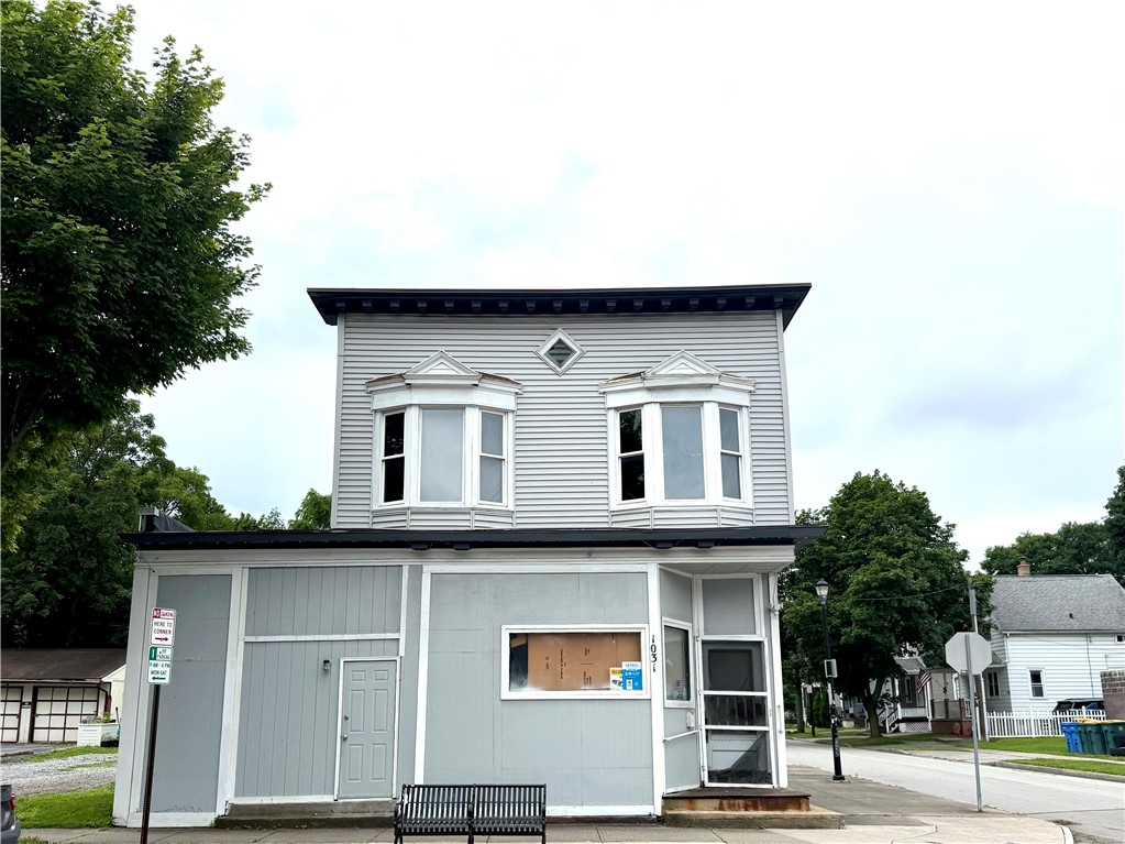 Two-story gray building with twin bay windows, a plywood-covered storefront window, and a sidewalk bench.