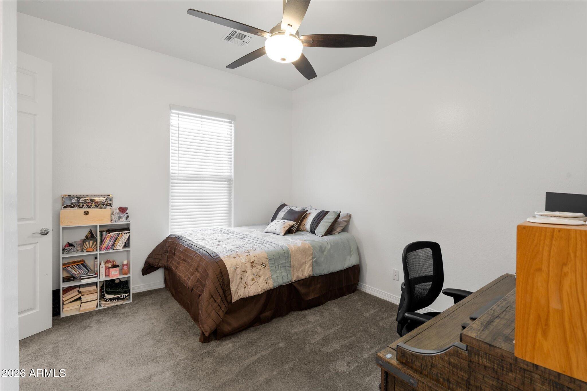 Bedroom with a patterned bed, window blinds, ceiling fan, and desk area.