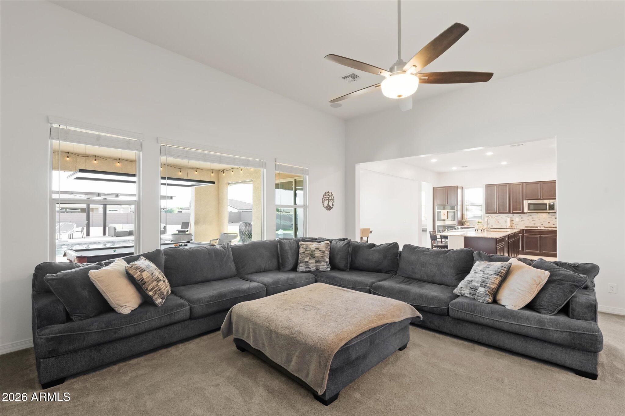 Furnished living room with a charcoal sectional, opening to a kitchen and windows overlooking a pool.