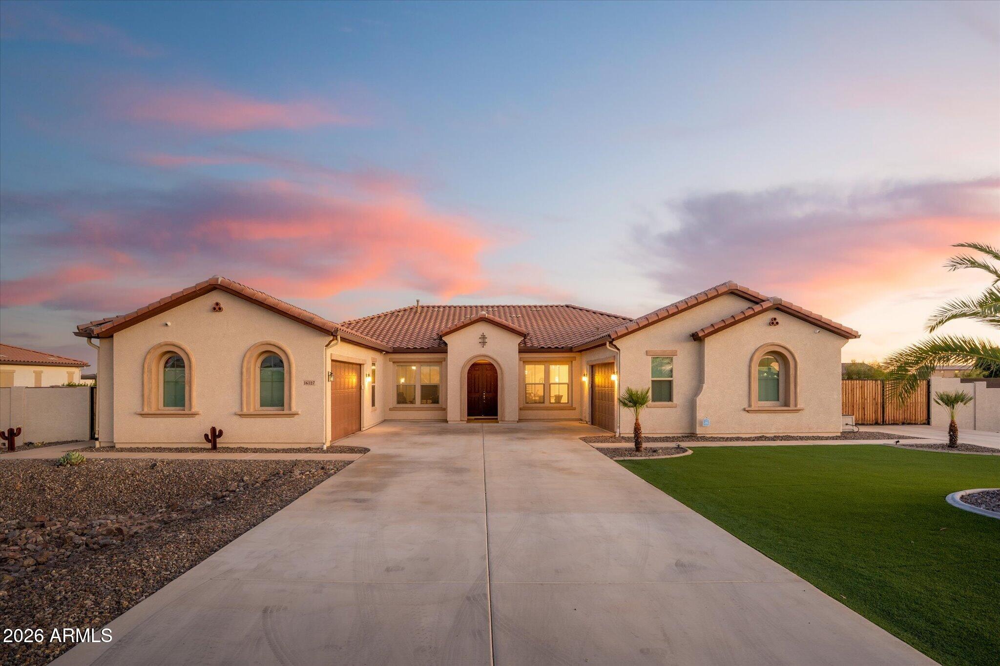 Single-story stucco home with arched windows, a wide driveway, and desert landscaping at dusk.