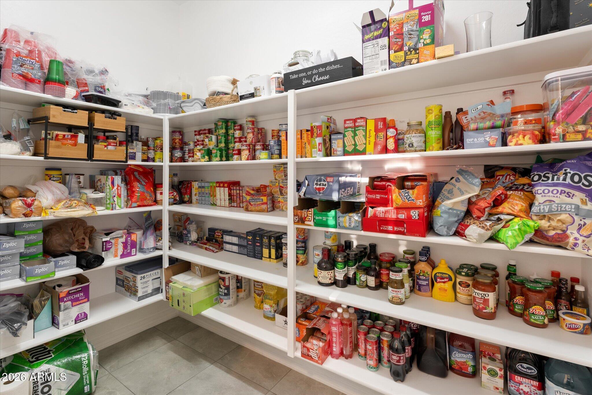 Walk-in pantry with wraparound shelves stocked with food, drinks, and household supplies.