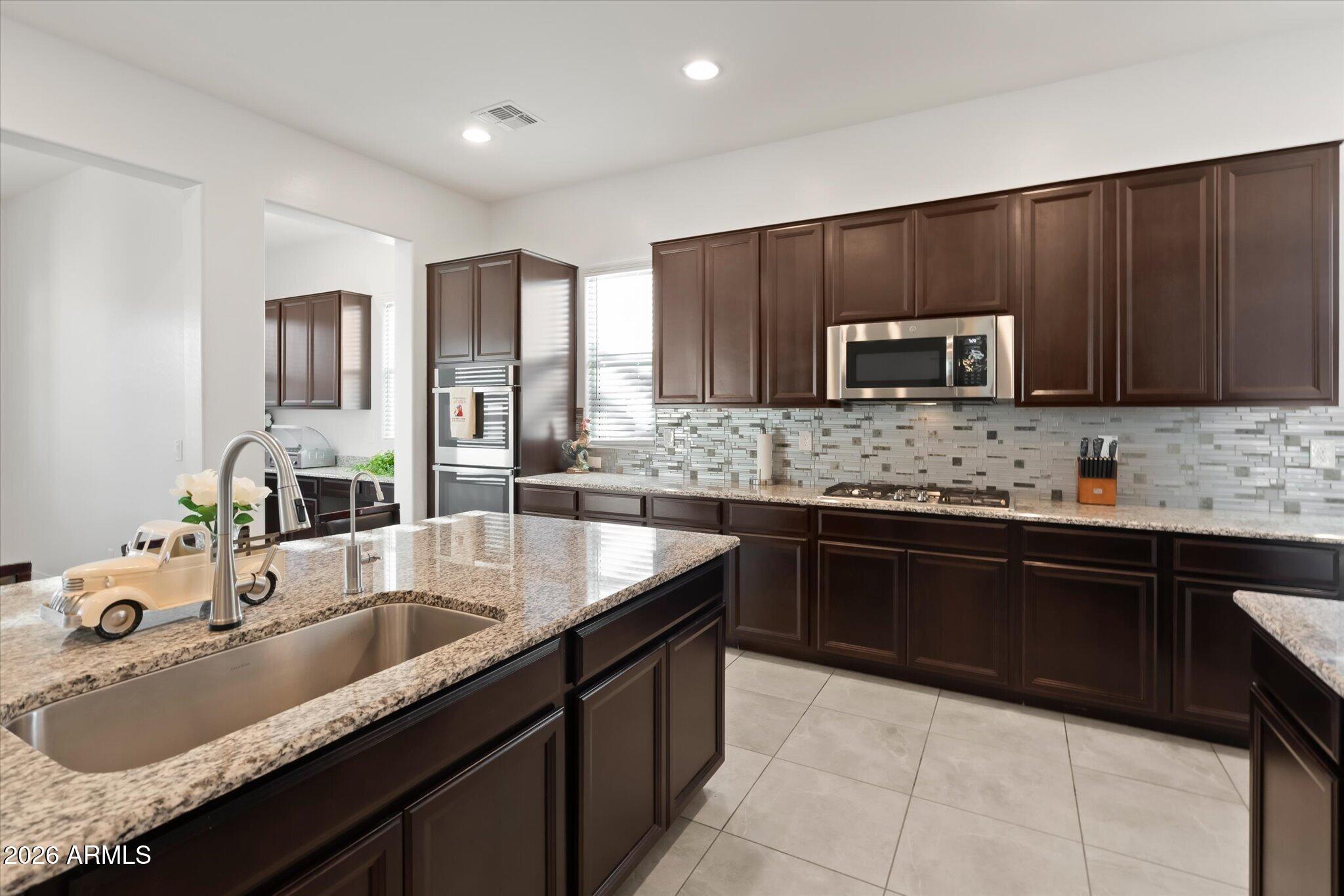 Kitchen with dark wood cabinets, granite island, deep sink, stainless appliances, and mosaic tile backsplash.