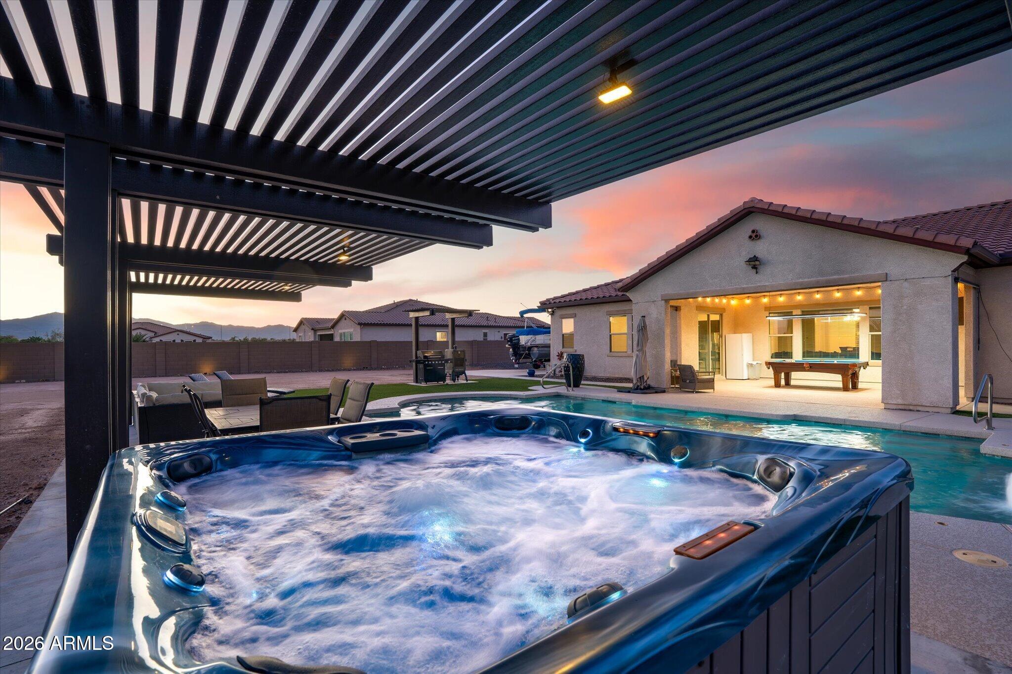 Backyard with a bubbling hot tub, swimming pool, and pergola at dusk.