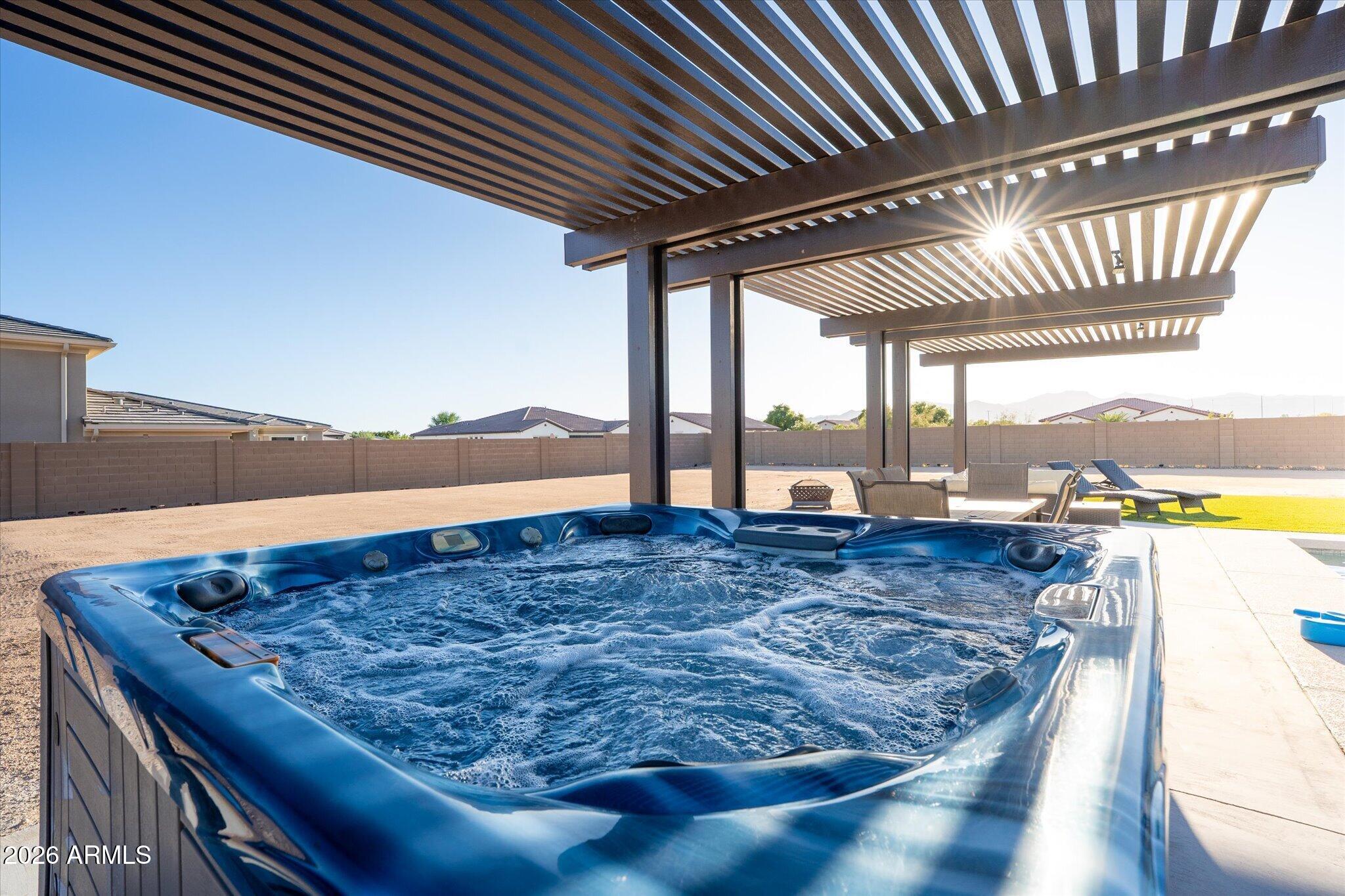Backyard hot tub beneath a slatted pergola, with patio furniture and neighboring homes beyond.