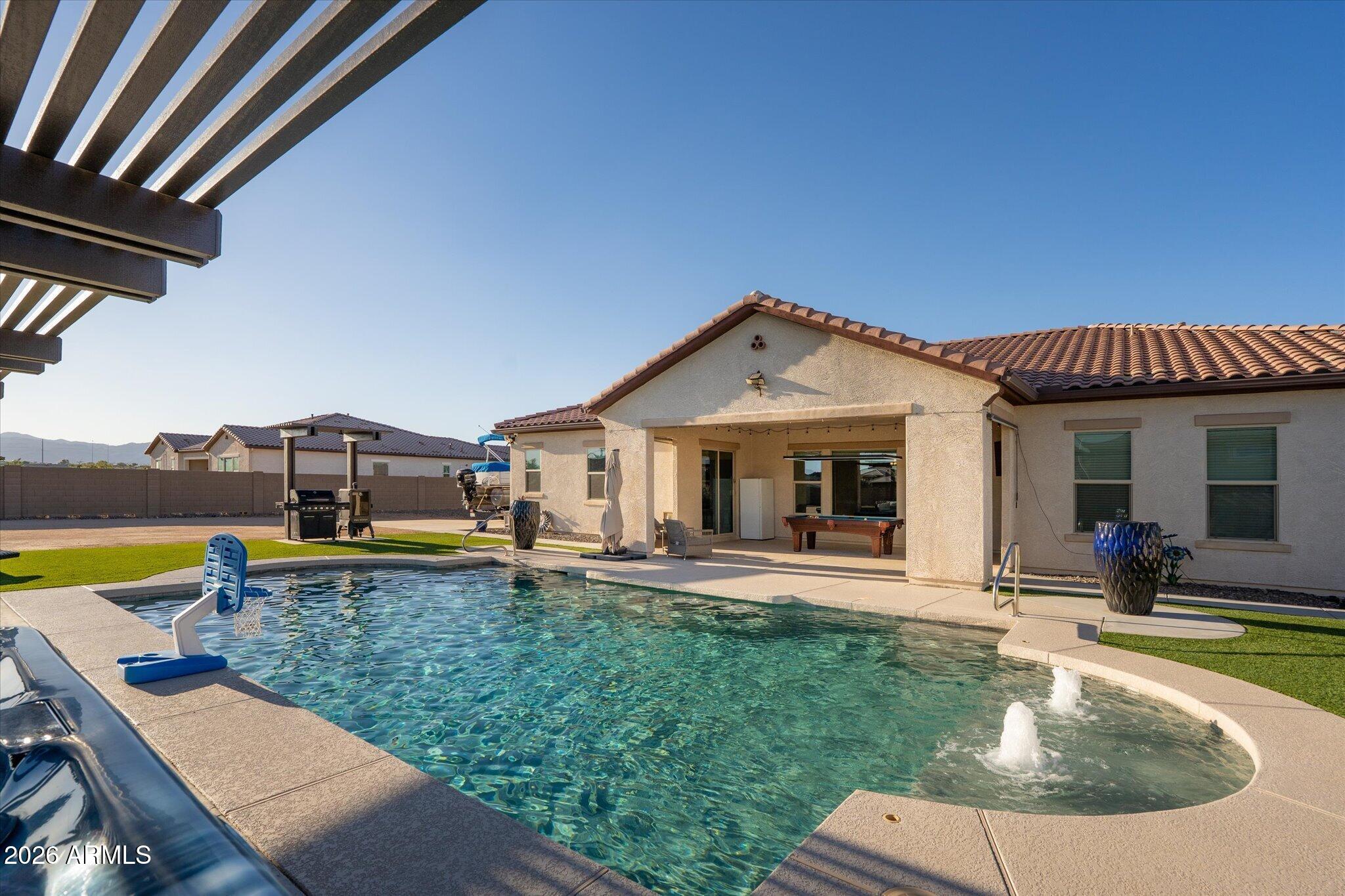 Backyard pool with fountains and basketball hoop beside a covered patio.