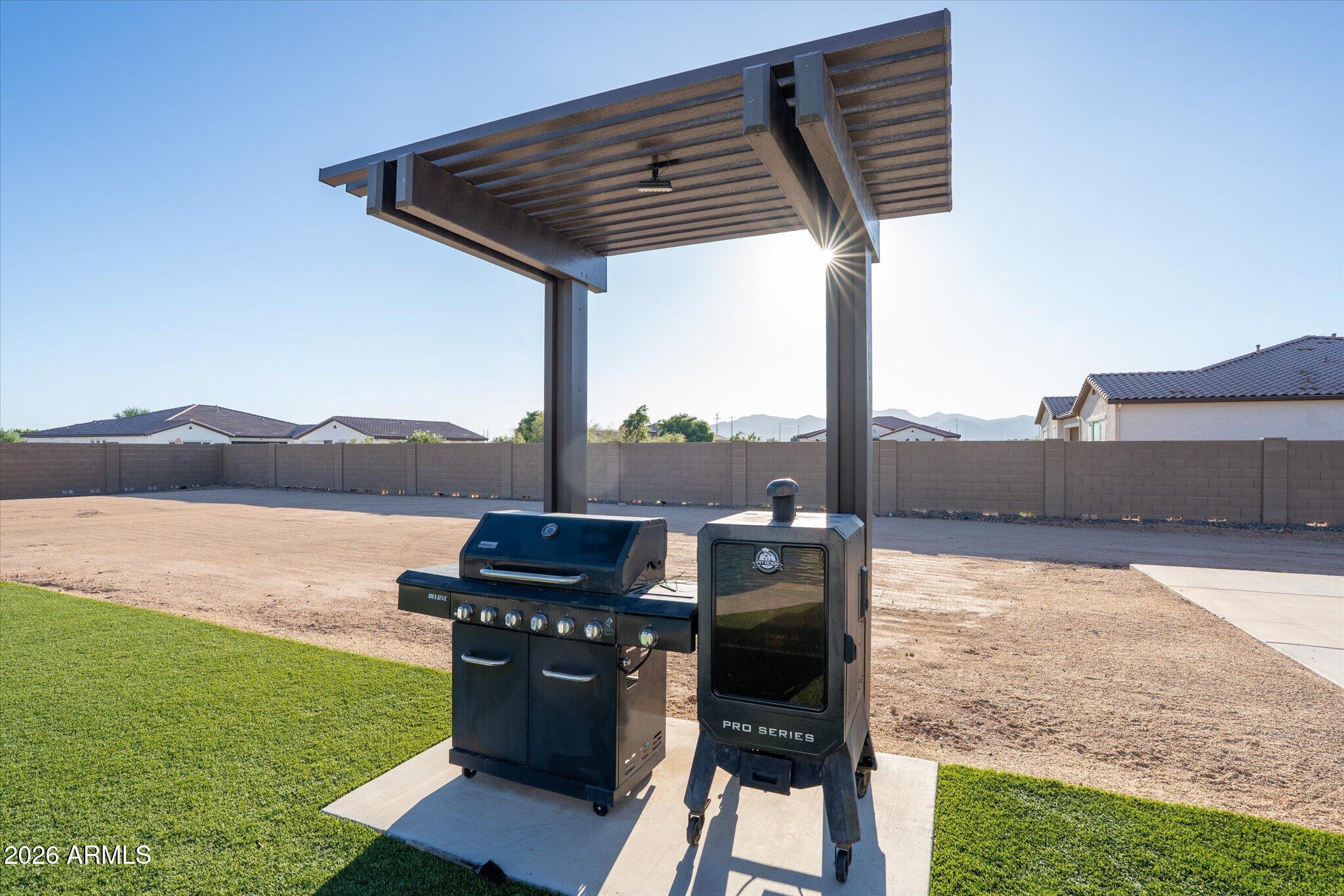 Backyard grill and smoker beneath a modern pergola, beside lawn and gravel yard.