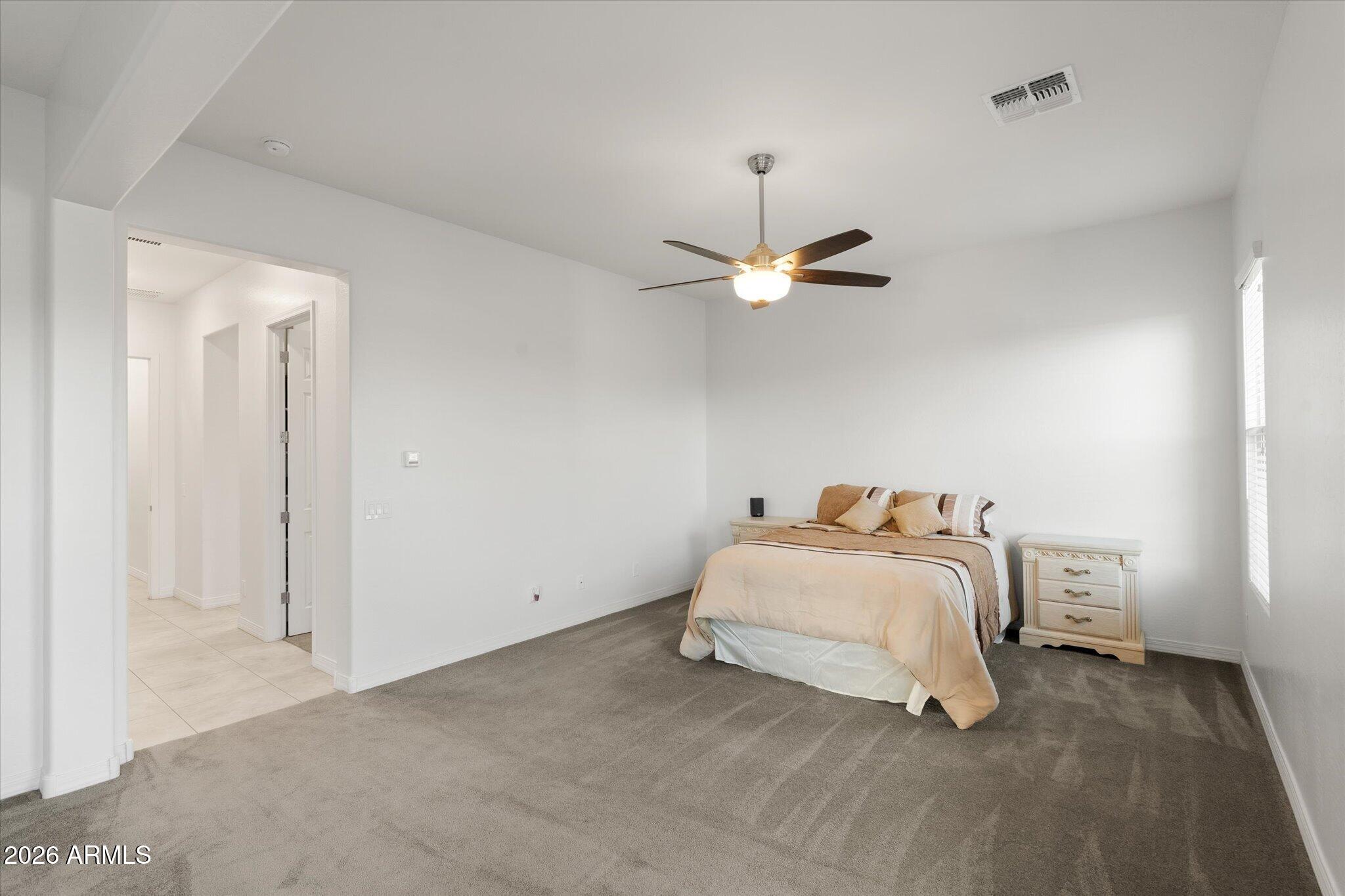 Furnished bedroom with beige bedding, gray carpet, and a ceiling fan with a lit fixture.