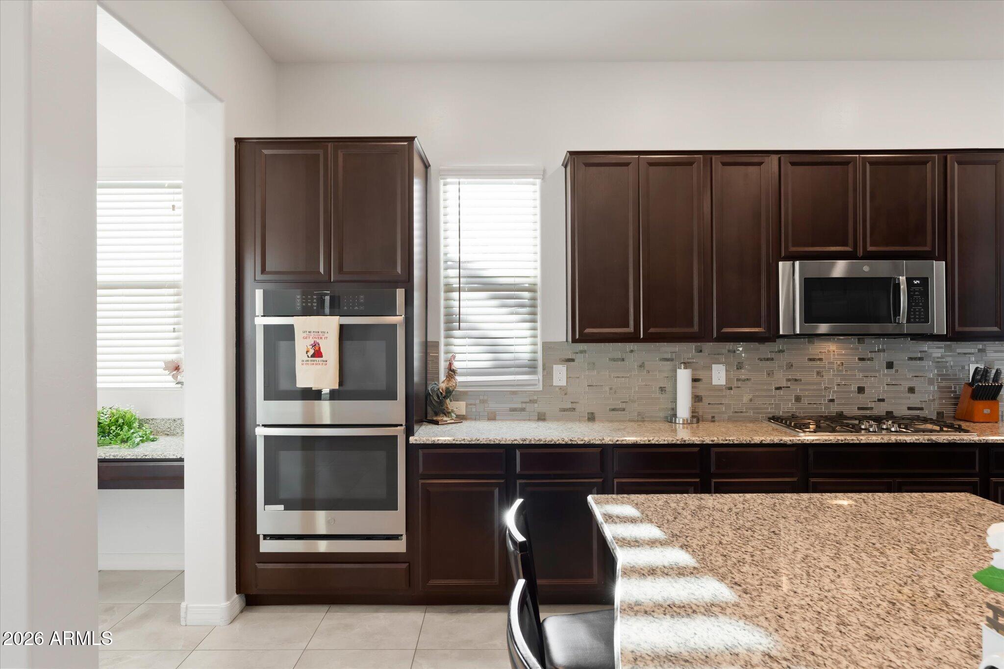 Kitchen with dark wood cabinets, stainless steel double ovens, mosaic backsplash, and a large granite island.