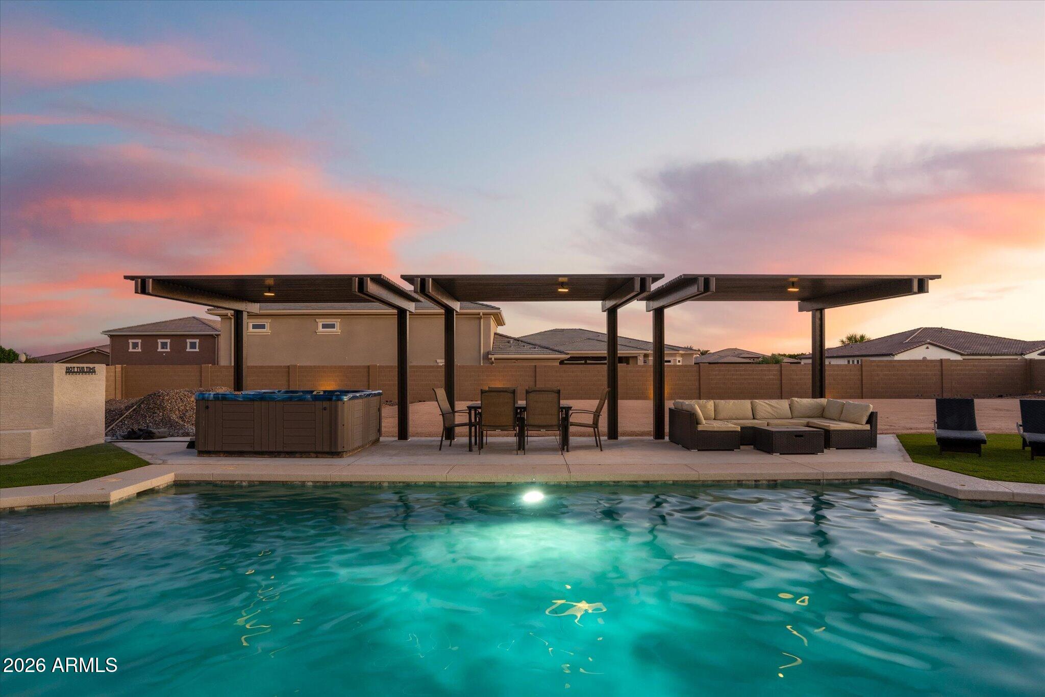 Backyard swimming pool with illuminated water, covered patio seating, and a hot tub at dusk.