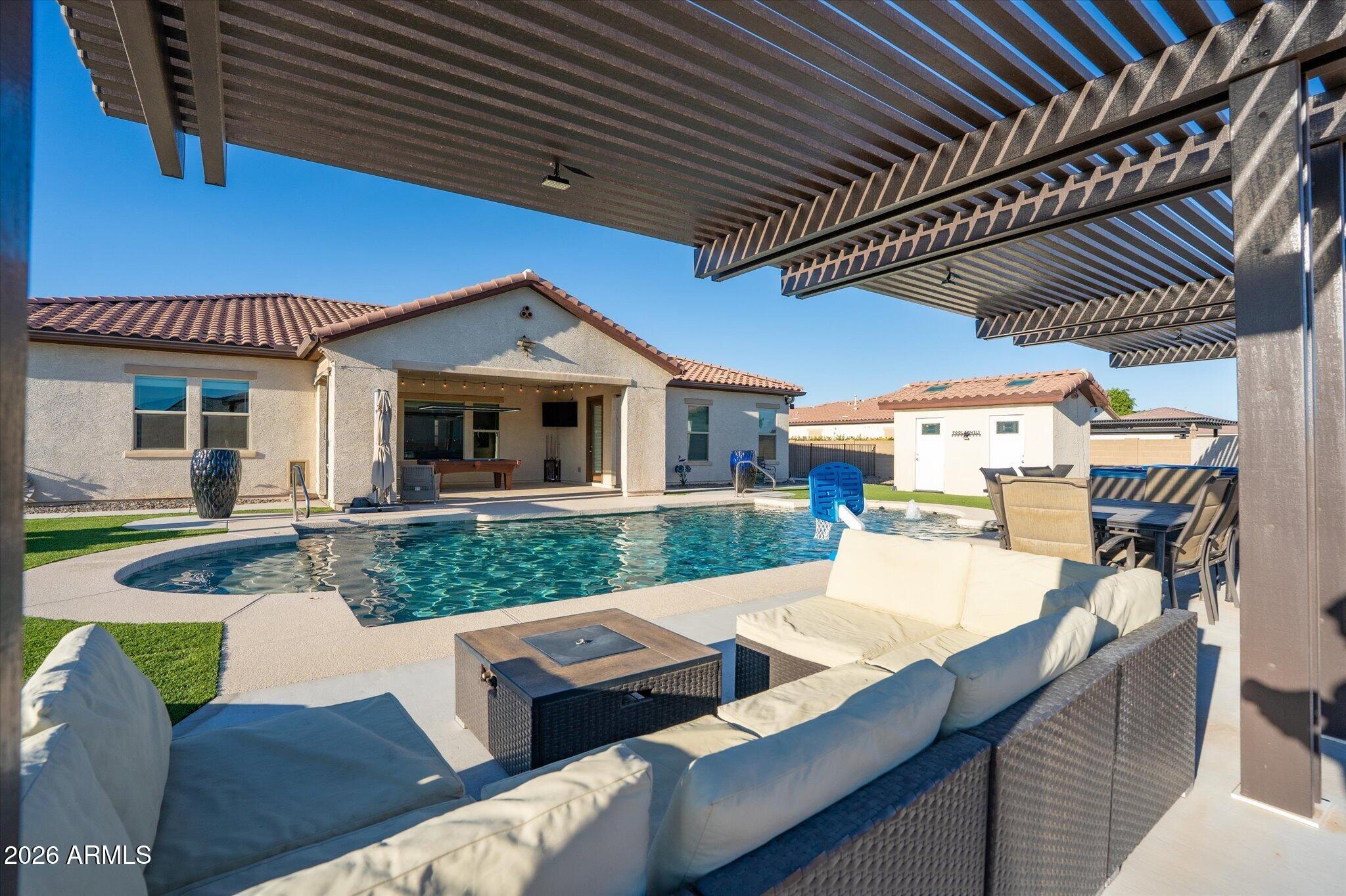 Backyard pool beneath a pergola, with outdoor lounge seating and a basketball hoop.