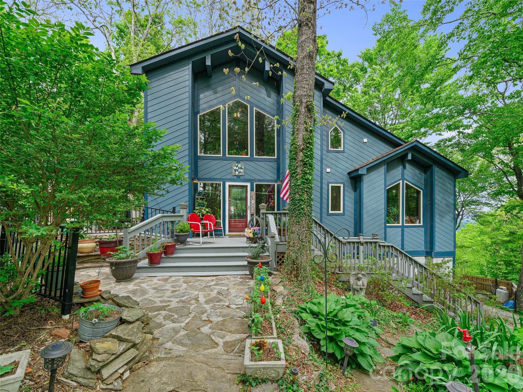 Blue house with a raised porch, red chairs, and a stone path through a leafy garden.