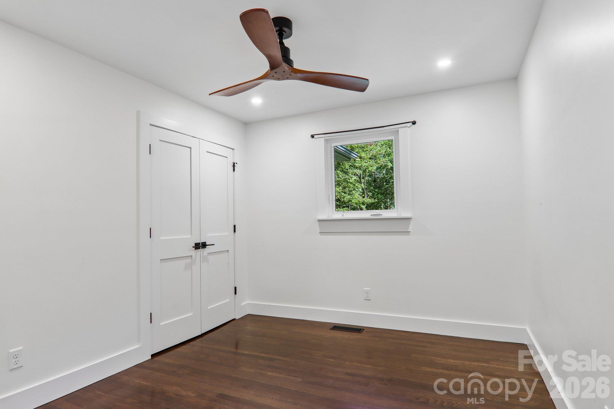 Empty bedroom with dark hardwood floors, double closet doors, and a wooden ceiling fan.