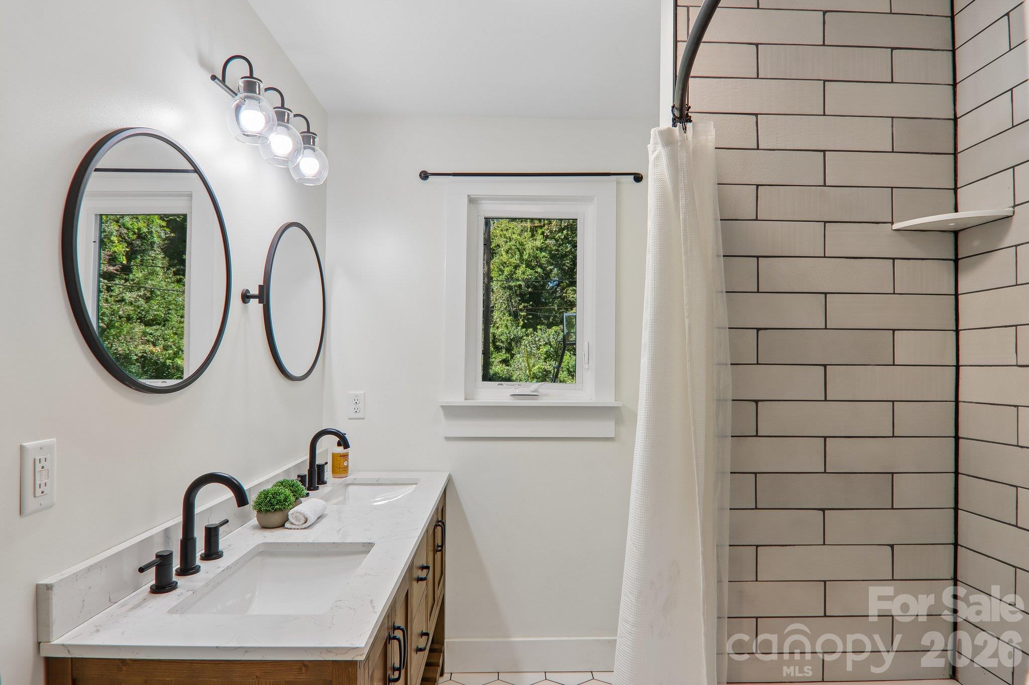 Bathroom with double vanity, oval mirrors, and a tiled shower beside a window.