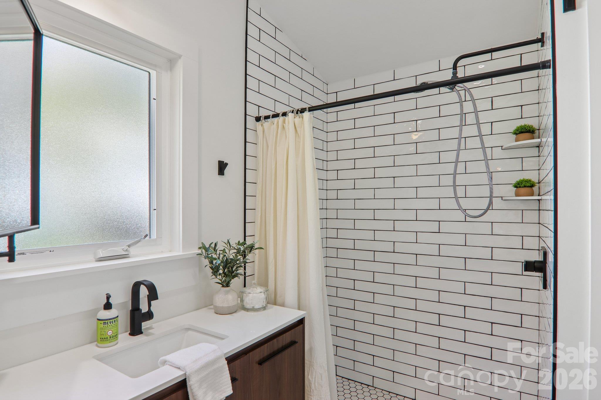 Bathroom with a white subway-tile shower, black fixtures, and a wood vanity beside a frosted window.