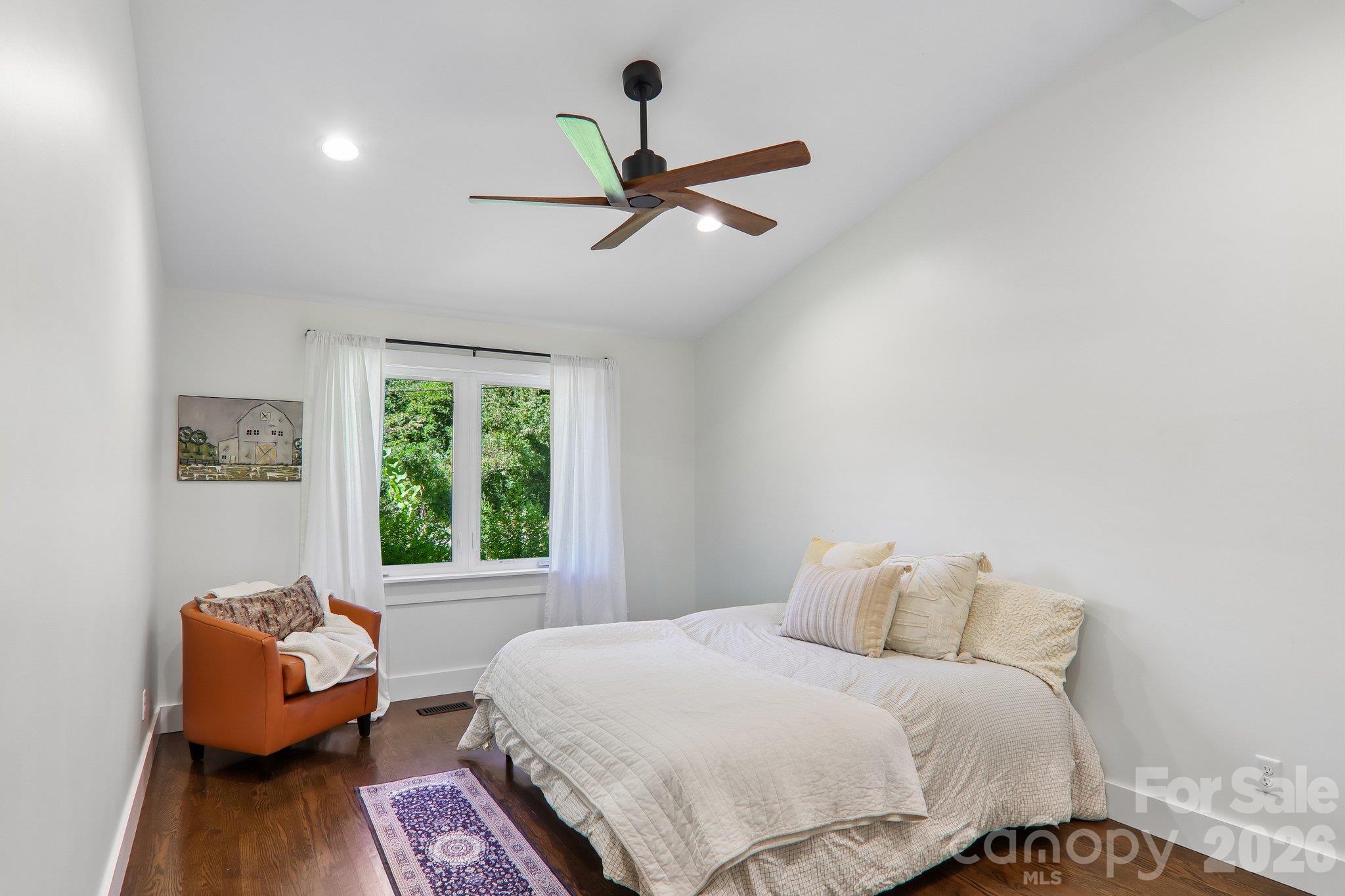 Furnished bedroom with a white bed, orange armchair, dark wood floor, and leafy window view.