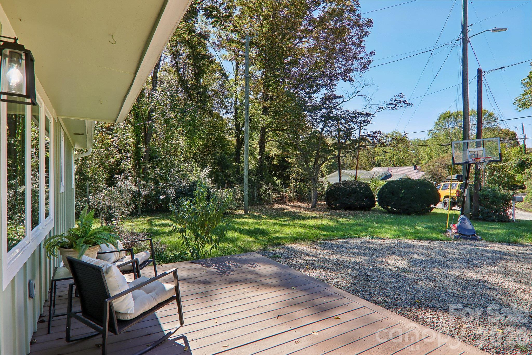 Covered wooden deck with cushioned seating overlooking a grassy, tree-lined yard and basketball hoop.