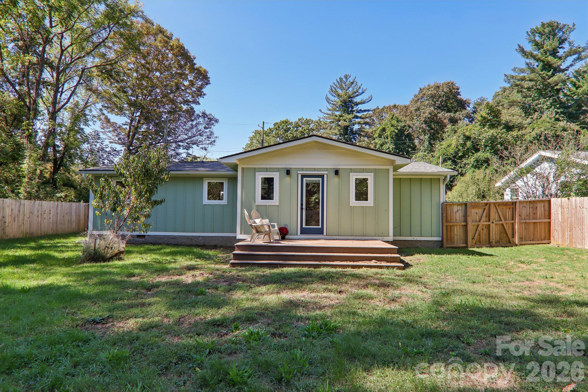 Green single-story home with a raised deck, chairs, fenced yard, and mature trees.