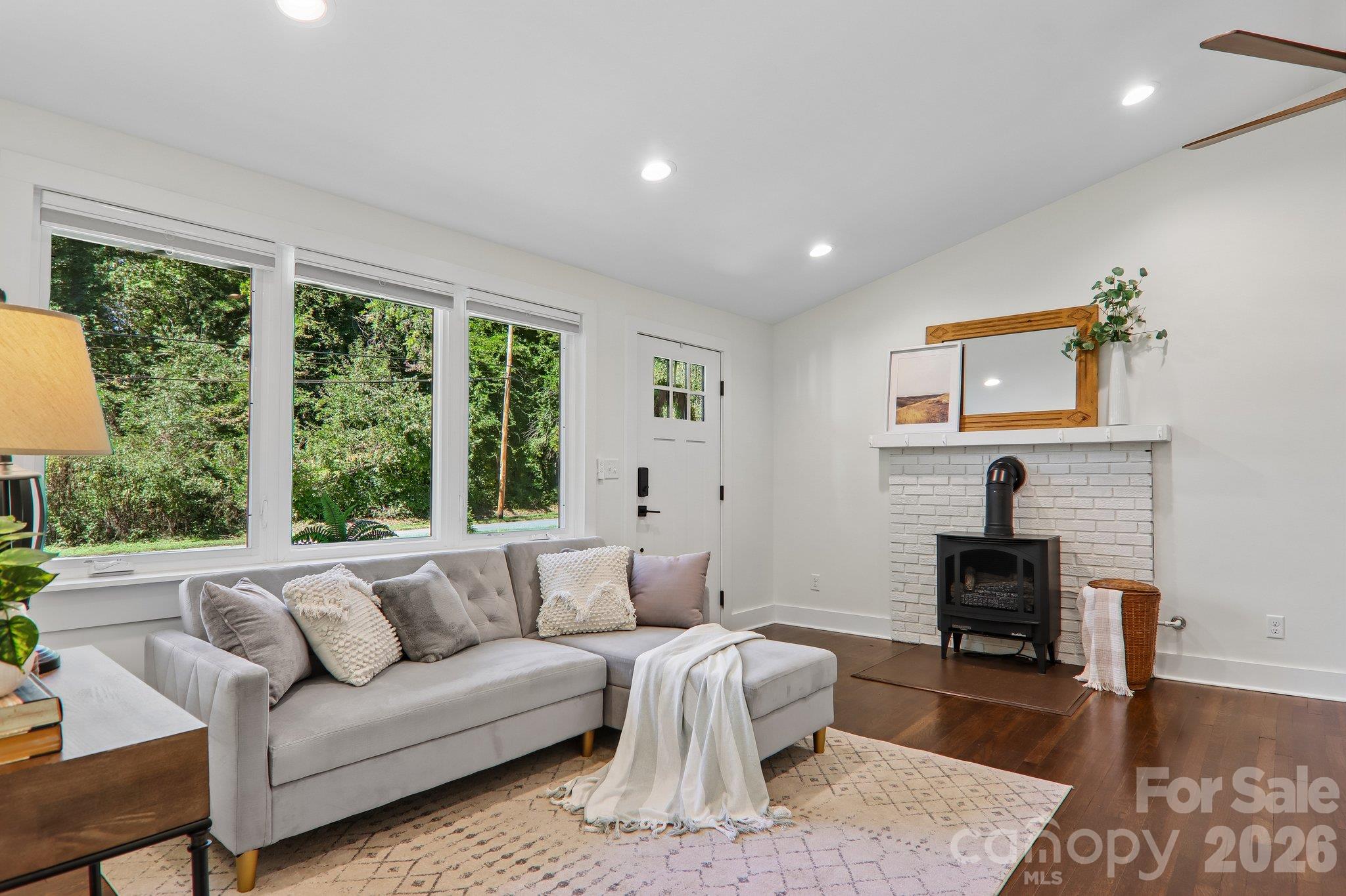 Staged living room with gray sectional, tree-filled windows, and a white brick fireplace with a black stove.