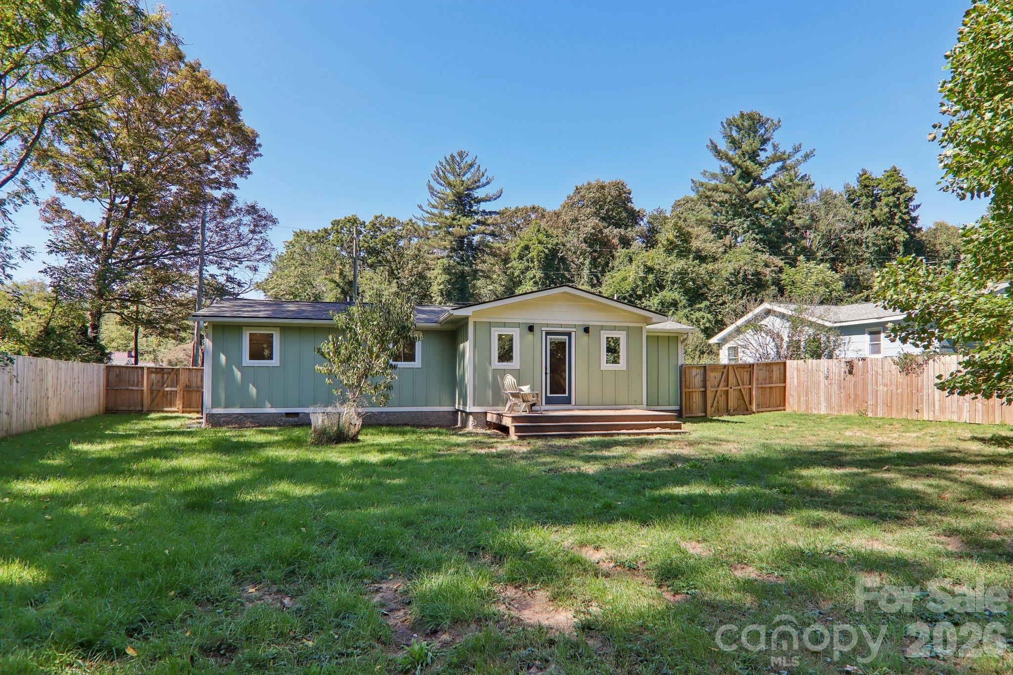 Sage-green single-story home with a small deck, grassy yard, and wooden privacy fence.
