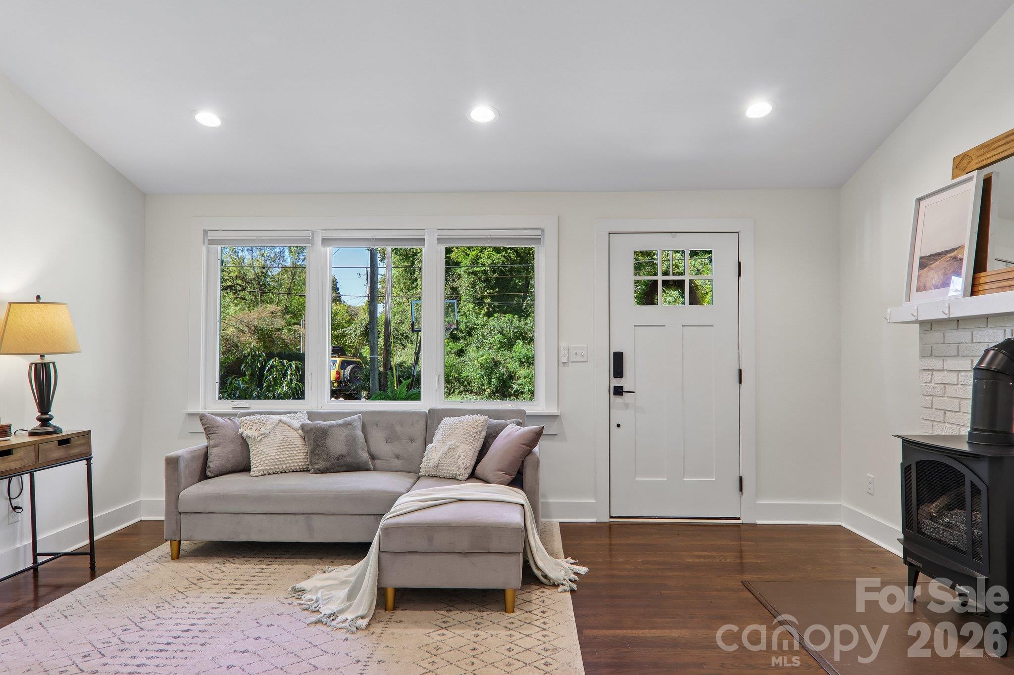 Living room with a gray sectional, wide windows, hardwood floors, and a black stove-style fireplace.