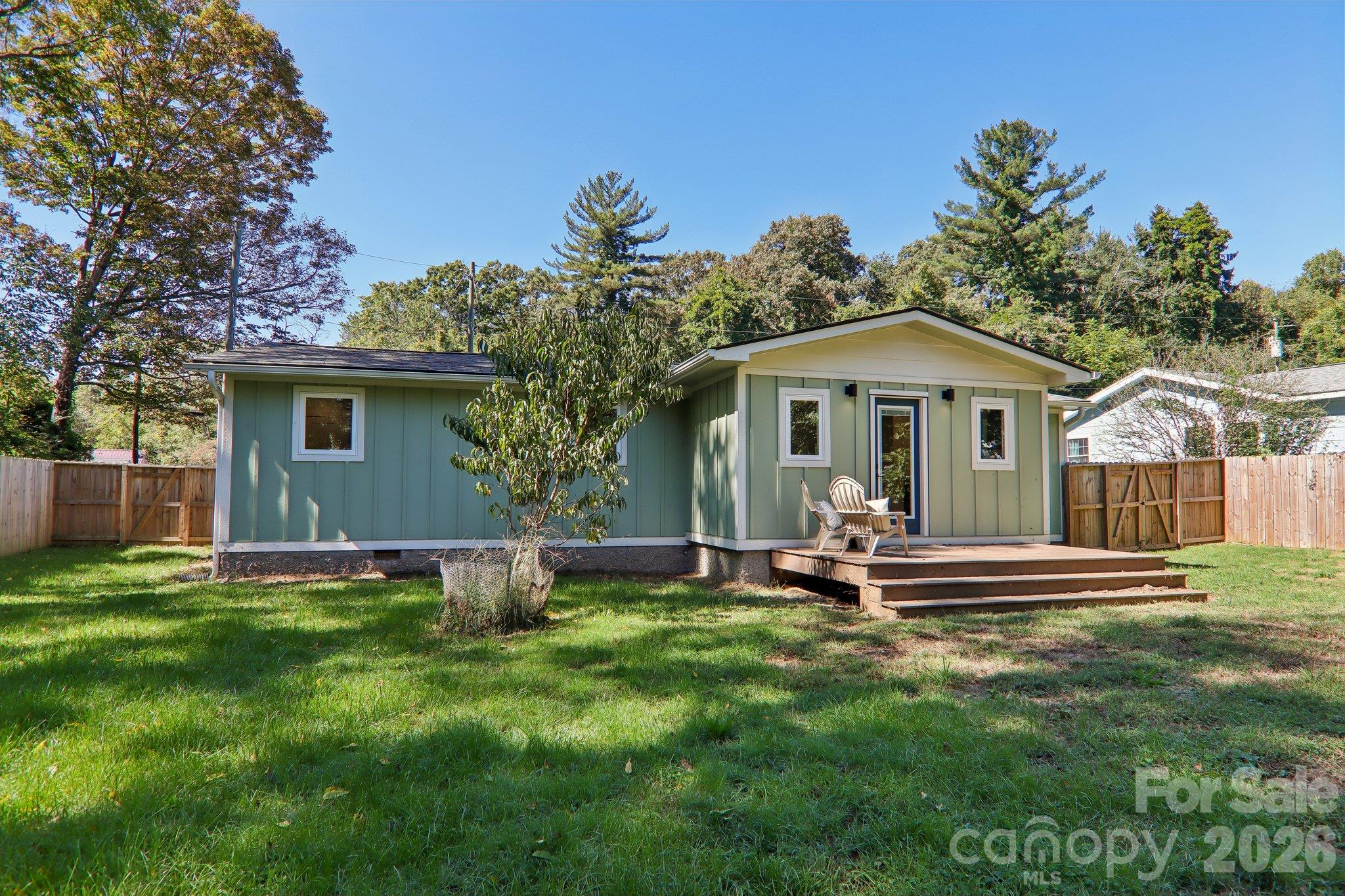 Green single-story home with a raised deck and chairs, set in a fenced grassy yard.