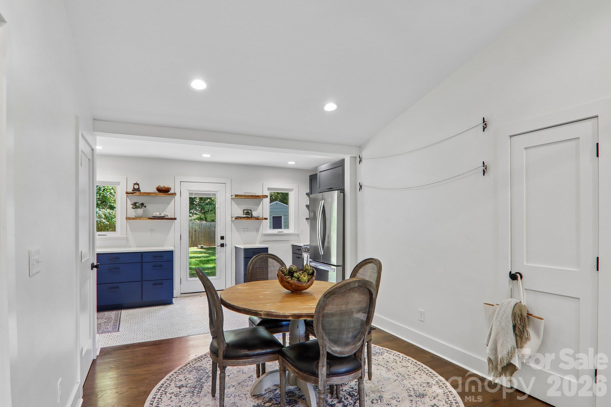 Dining area with a round wooden table beside a bright kitchen with navy cabinets and stainless steel appliances.
