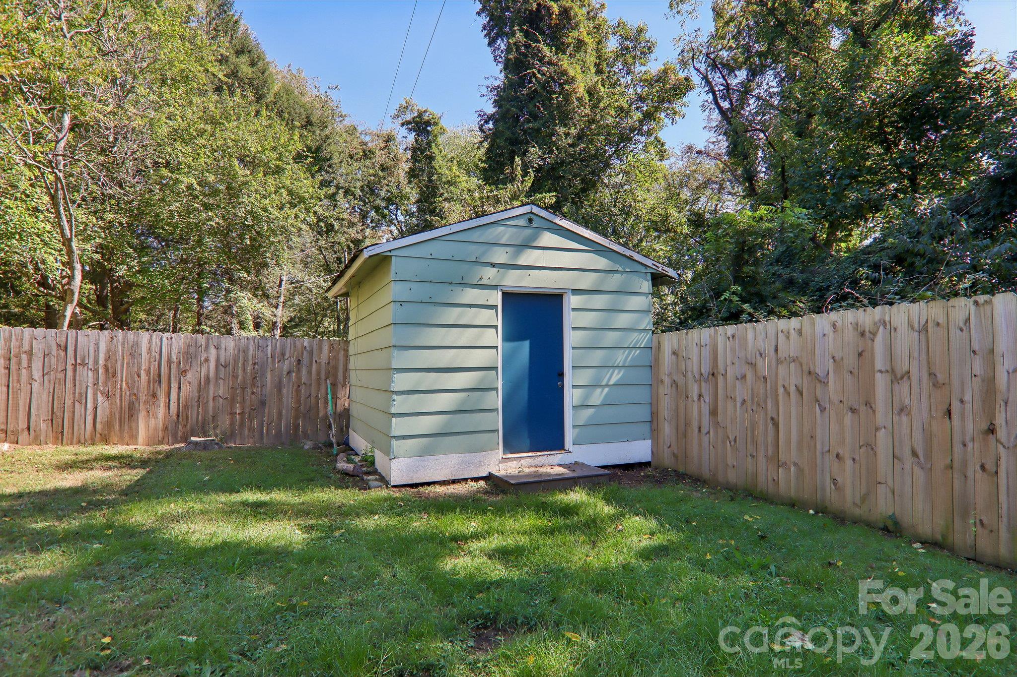 Pale green backyard shed with a blue door, surrounded by a wooden fence and trees.