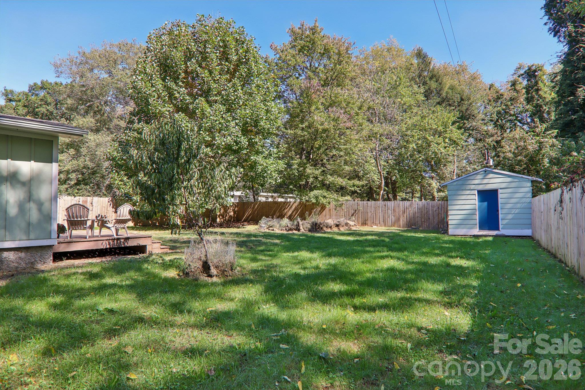 Fenced backyard with a broad lawn, mature trees, a small deck, and a detached shed.